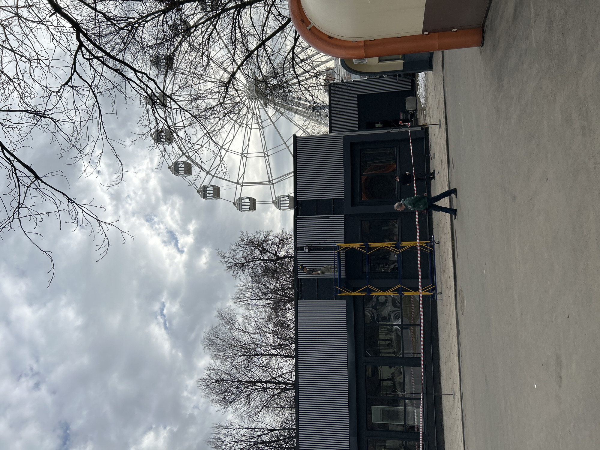 The image shows a ferris wheel behind a modern building with scaffolding. Two people are visible, one walking past the building. Bare trees are visible on the left side of the image. The sky is overcast with clouds. The location is the VDNKh park in Moscow, Russia.