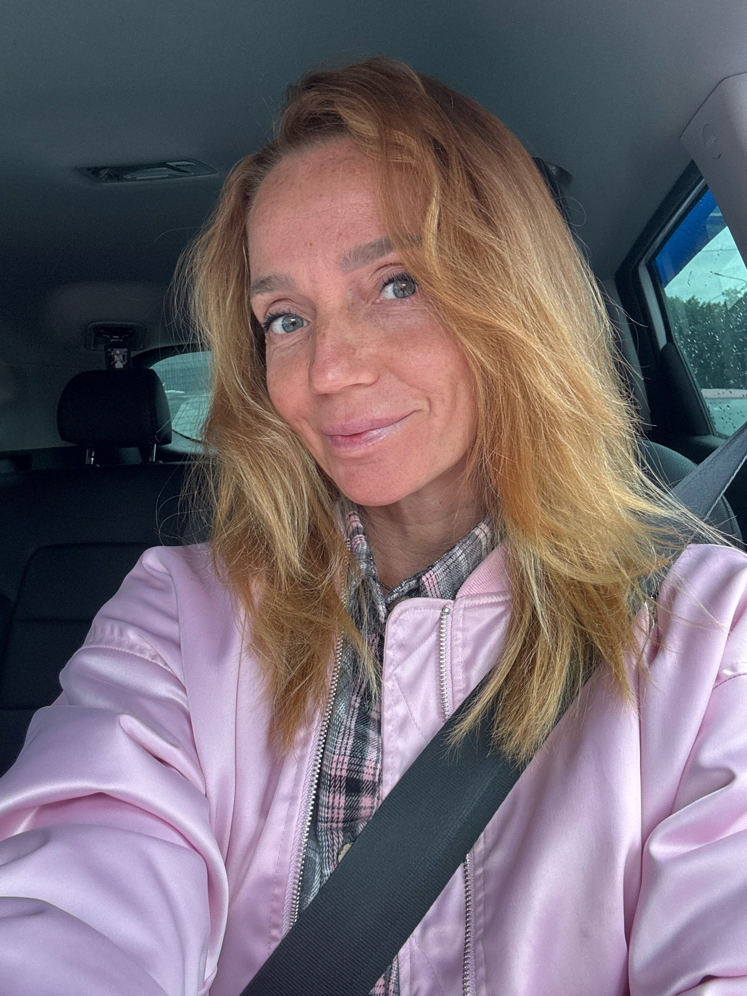 A medium shot portrait of a woman with blonde hair and blue eyes, wearing a pink bomber jacket over a plaid shirt, is shown. She is sitting in the driver's seat of a car, with a seatbelt across her chest. The background shows the car's interior and a rainy day outside the window. The woman is looking at the camera with a slight smile. The location is not specified in the image.
