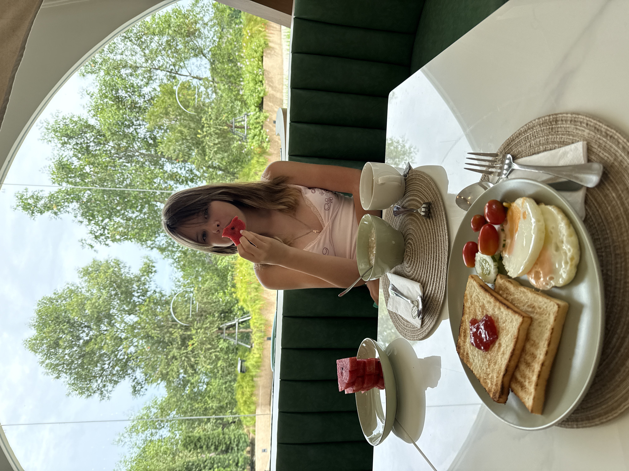 A young Caucasian girl with brown hair is seated at a white table in a restaurant. She is wearing a pink camisole. In front of her, a breakfast is laid out: two slices of toast topped with jam, two fried eggs, cherry tomatoes, and cucumber slices on a green plate. To her left, a bowl of oatmeal, a cup of tea, and a plate of sliced watermelon are also present. The girl is holding a slice of watermelon to her mouth, mid-bite. The restaurant features green banquette seating and a large arched window that overlooks lush green trees. The establishment is located in Khok Kham Subdistrict, Thailand.