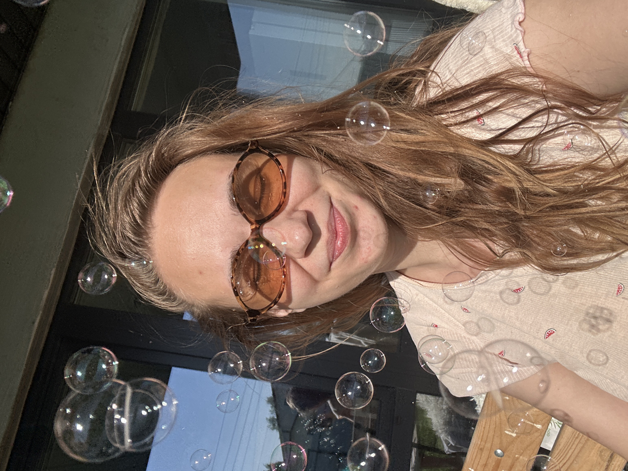 A woman with light brown hair and sunglasses is shown in a close-up shot, with numerous soap bubbles floating around her. The woman is wearing a light-colored shirt with small watermelon designs. The background includes a window and what appears to be outdoor vegetation. There is no clear indication of the city of Kharnavy, Russia in the image.
