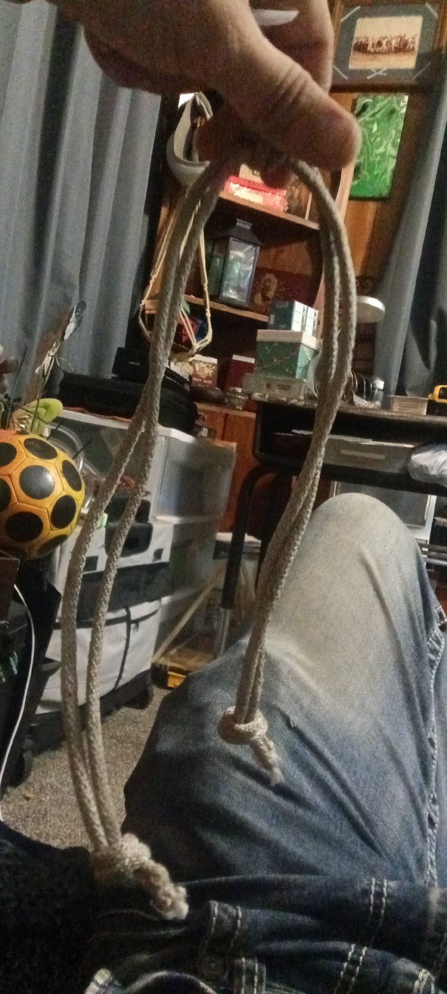 In Coal Hill, United States of America, an image captures a close-up view of a person's hand holding a rope. The rope, made of a braided, light-colored material, forms a loop with two knotted ends. The rope is suspended in front of a background that includes blue curtains, shelves filled with various items such as boxes and framed pictures, a black electronic device, and a yellow and black ball. The lower portion of the image shows the person's legs, clad in denim jeans. The scene appears to be indoors, possibly a living room or storage area, with a carpeted floor.