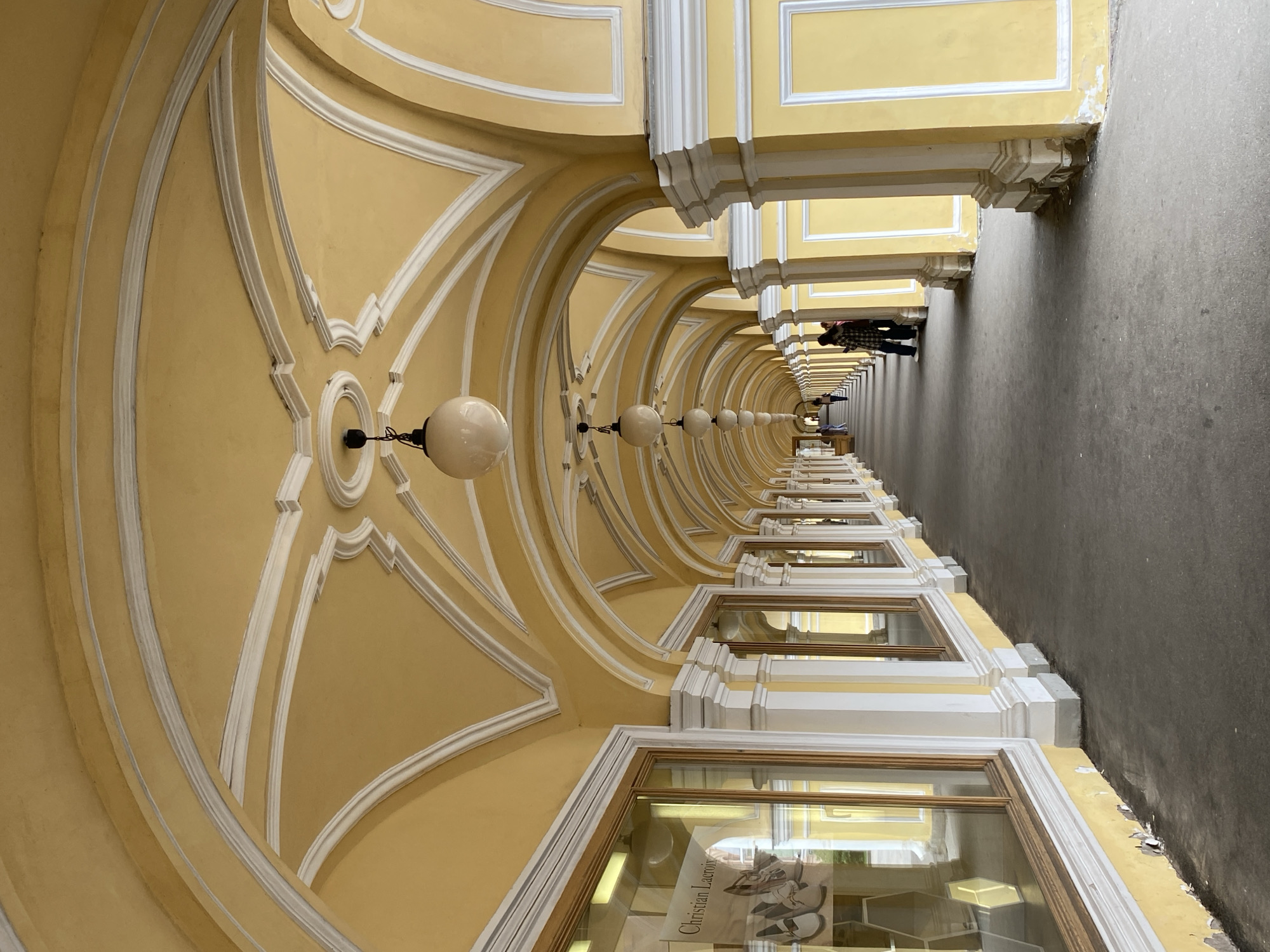 The image depicts an interior view of a long, covered arcade in Saint Petersburg, Russia. The architecture features a repeating pattern of arched ceilings painted yellow with white decorative trim. Hanging spherical white lights are spaced along the arcade. On the left side, there are shop windows with displays, one featuring a sign that reads "Christian Lacroix." On the right side, the arcade is supported by columns, also painted yellow with white trim, and there are recessed panels with white borders. The floor is paved with dark asphalt. In the distance, several people are walking along the arcade, and two individuals are standing on the right side, facing away from the camera. The overall impression is one of a grand, classical architectural space, likely a public building or promenade.