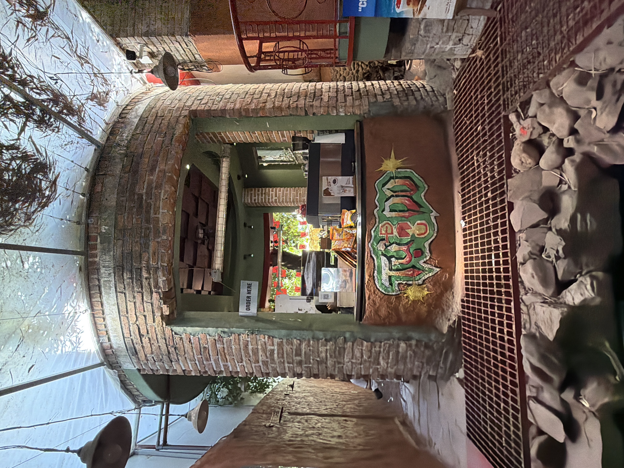 The image shows an outdoor cafe counter in Nha Trang Ward, Vietnam. The counter is made of brick and has a circular design. There is a coffee machine and various snacks displayed on the counter. A sign above the counter reads "ORDER HERE". The cafe appears to be situated in a narrow alleyway with a grated floor in the foreground. In the background, there is a red railing and some greenery. The ceiling is covered with a translucent material, possibly plastic sheeting, with dried leaves or branches on top, allowing diffused sunlight to filter through. The overall ambiance suggests a rustic and somewhat bohemian setting.