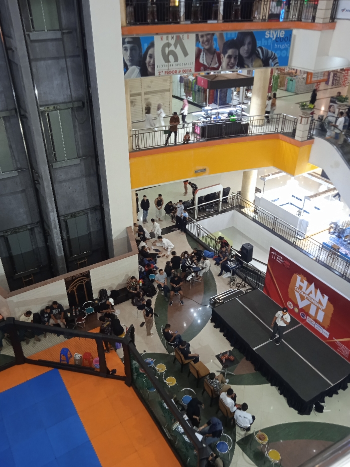 The image shows a scene inside a shopping mall in Surakarta, Indonesia, located at coordinates -7.567 latitude and 110.807 longitude. There are people sitting and standing around a stage set up in the middle of the mall, facing a large banner that reads "HAN VI CHAMPIONSHIP." There are some people sitting on benches in the background. There are multiple levels to the mall, with an elevator in the left corner of the image. The scene is brightly lit, with a mix of dark gray and orange tones. There is a blue and orange mat in the foreground.