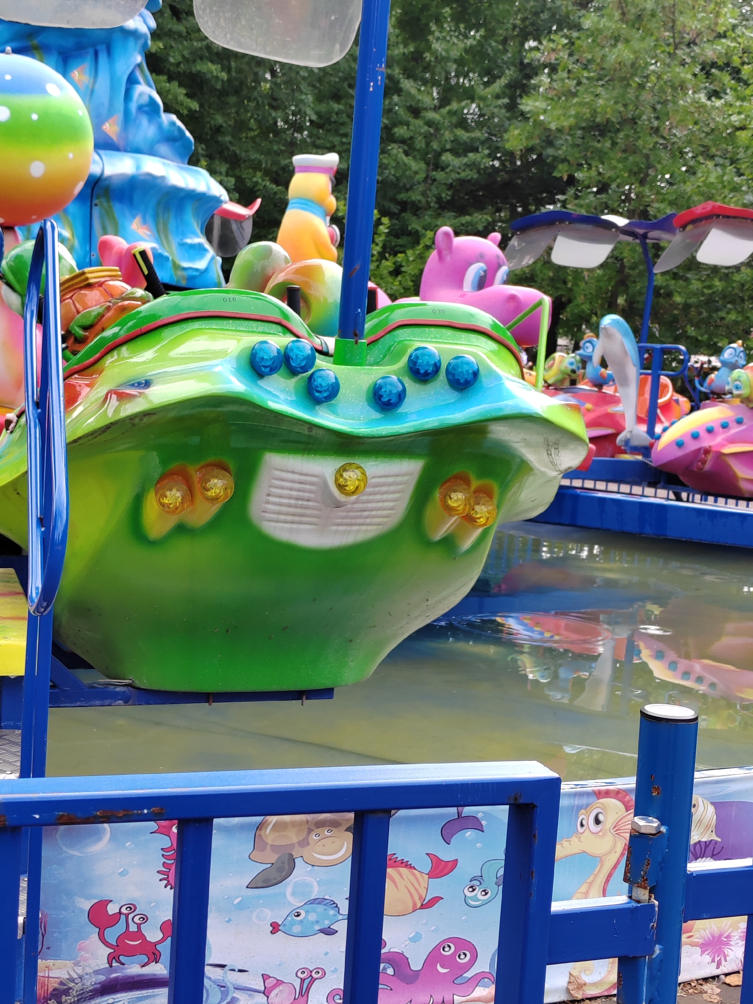 This is a close-up, eye-level, medium shot of a colorful amusement park ride, specifically a boat-themed ride, in Brest, Belarus. The ride is predominantly green and blue, with several cartoonish sea creatures adorning its exterior. In the foreground, a blue metal railing with some rust visible separates the viewer from the ride. Behind the railing, a blue barrier is decorated with cartoon images of sea life, including a red crab, a blue fish, a turtle, and a pink octopus. The boat ride itself is a vibrant green, with yellow and blue lights scattered across its surface. Reflections of the ride and the surrounding greenery are visible in the shallow water at the base of the attraction. In the background, lush green trees suggest the park is situated in a natural, outdoor environment. The overall atmosphere is playful and lighthearted, indicative of a family-friendly amusement park.
