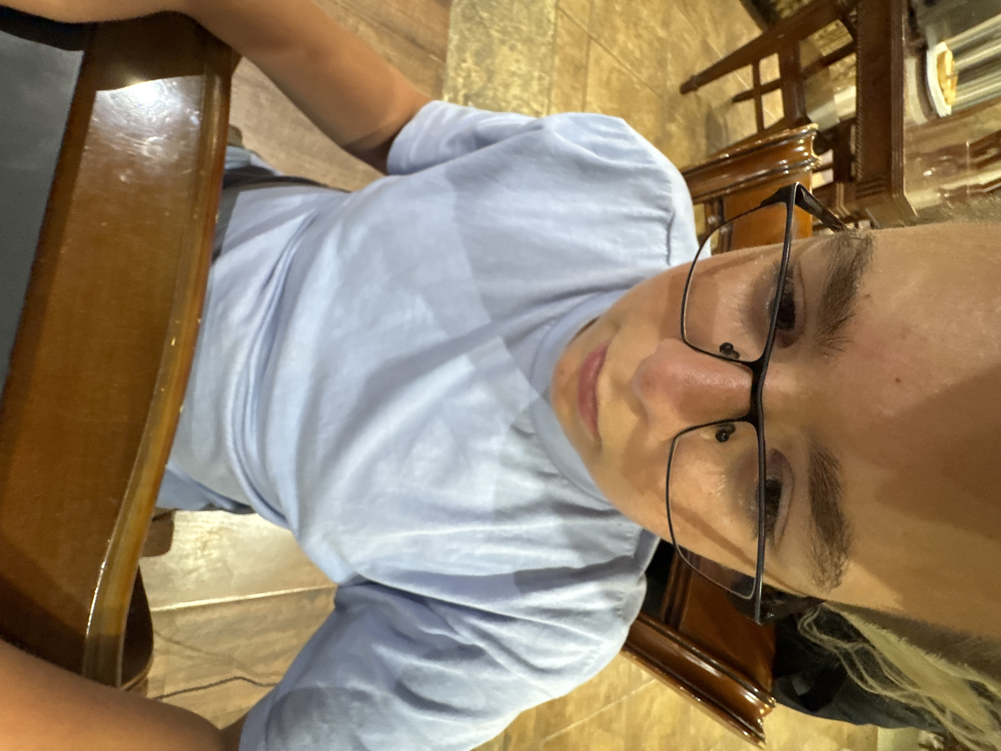 The image shows a person wearing glasses and a light blue t-shirt. The person is seated at a wooden table, which appears to be in a restaurant or dining area, suggested by the presence of other wooden furniture in the background and the tiled flooring. The lighting in the image is warm, indicating it might be evening or indoors. The person's face is partially visible, with their gaze directed away from the camera. The scene appears casual and relaxed. There is no visible text in the image.