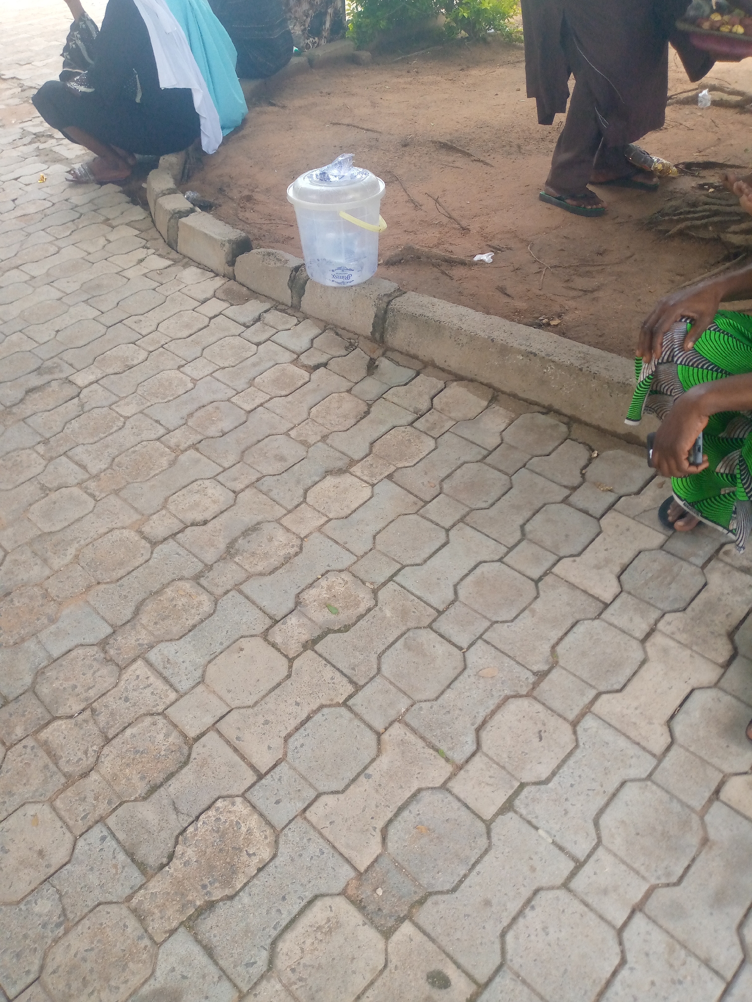 A group of individuals are gathered outdoors on a dusty ground next to a paved walkway in Jalingo, Nigeria. In the foreground, a clear plastic bucket with a yellow handle sits on a concrete curb. The bucket appears to contain water and possibly some debris.
To the left, a person wearing a black garment is crouched on the paved walkway. They are partially covered by light blue and white fabric. To the right of the bucket, a person is seated, wearing a green and black patterned garment and dark sandals. Their hands are visible, and they appear to be holding a dark object, possibly a phone. Further in the background, another individual is standing, wearing a dark brown robe and dark sandals.
The environment is characterized by dry, sandy soil with scattered leaves and debris. The paved walkway is made of interlocking concrete blocks, worn and dusty. The overall scene suggests a casual gathering or an area of public access in Jalingo.