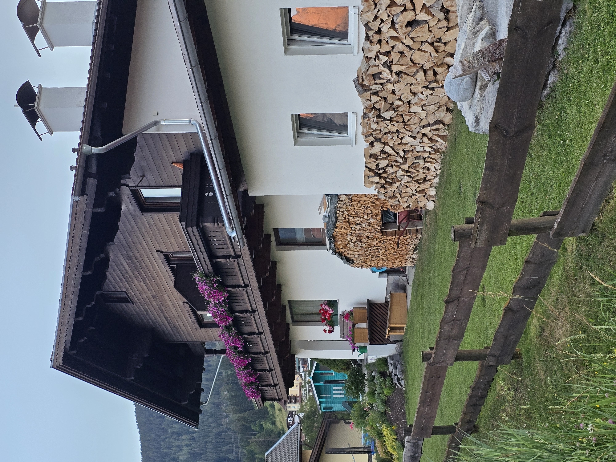 The image depicts a traditional Austrian house in Lermoos, Austria, characterized by its dark wood siding, white stucco walls, and a steeply pitched roof with decorative eaves. A wooden balcony, adorned with vibrant pink flowers, extends across the upper level. Two white chimneys with dark caps rise from the roofline. In front of the house, a substantial pile of neatly stacked firewood is visible, along with a smaller, partially covered pile. The ground floor features several windows, some reflecting the warm orange hues of the setting sun. A lush green lawn surrounds the house, bordered by a rustic wooden fence. Various garden elements are present, including flower beds, planters, and a green watering can. In the background, a dense forest covers a hillside, with a glimpse of other buildings in the distance. The overall scene conveys a sense of rural tranquility and traditional alpine living.
