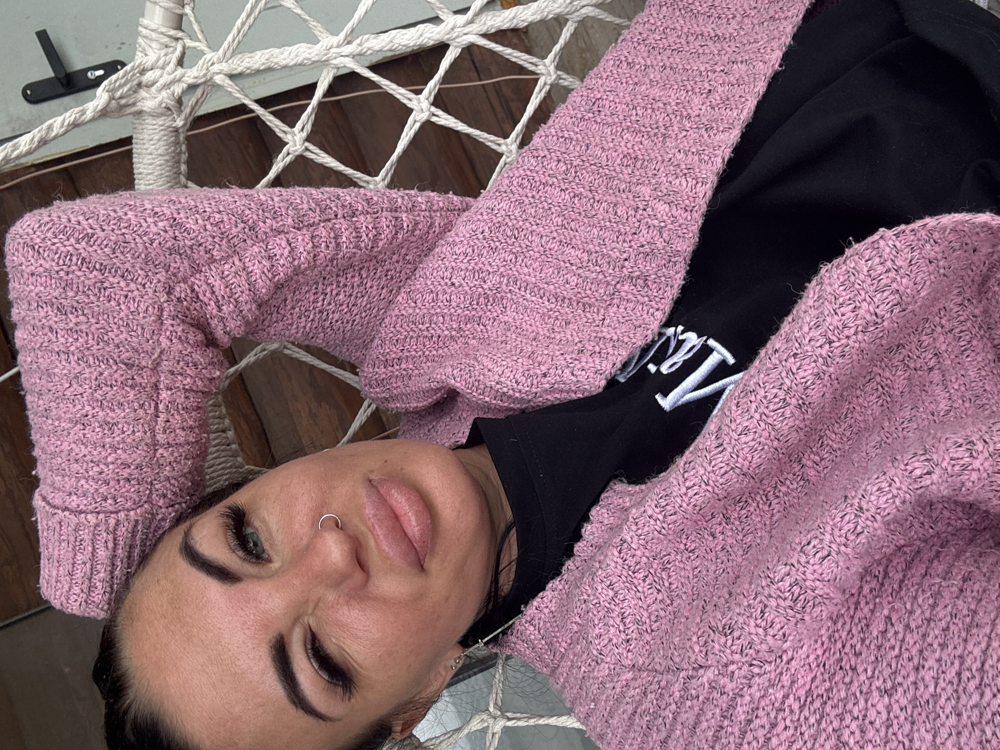 A woman is lying on a hammock, wearing a pink knitted cardigan and a black t-shirt. She is looking at the camera with one eye closed. The hammock is made of white rope and is suspended from a white pole. The background shows wooden panels and a door handle. The image is taken in a close-up shot, focusing on the woman's face and upper body. It is not possible to determine the specific location in Северянин Снт, Russia from the image.