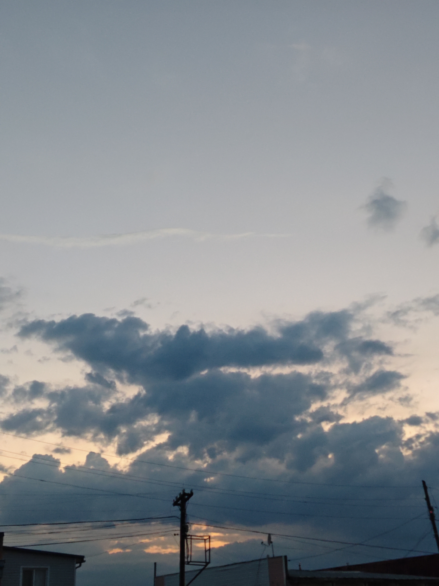 The image presents a view of a cloudy sky, likely captured around sunset or sunrise, over a cityscape in what appears to be Фоки, Russia. The sky transitions from a light gray-blue at the top to a warmer, peach-colored hue near the horizon. Clouds of varying density and dark gray color dominate the middle portion of the frame. Below the clouds, silhouetted rooftops and building structures are visible, along with utility poles and wires. The overall environment suggests an urban or suburban setting. There is no indication of people in this specific image.