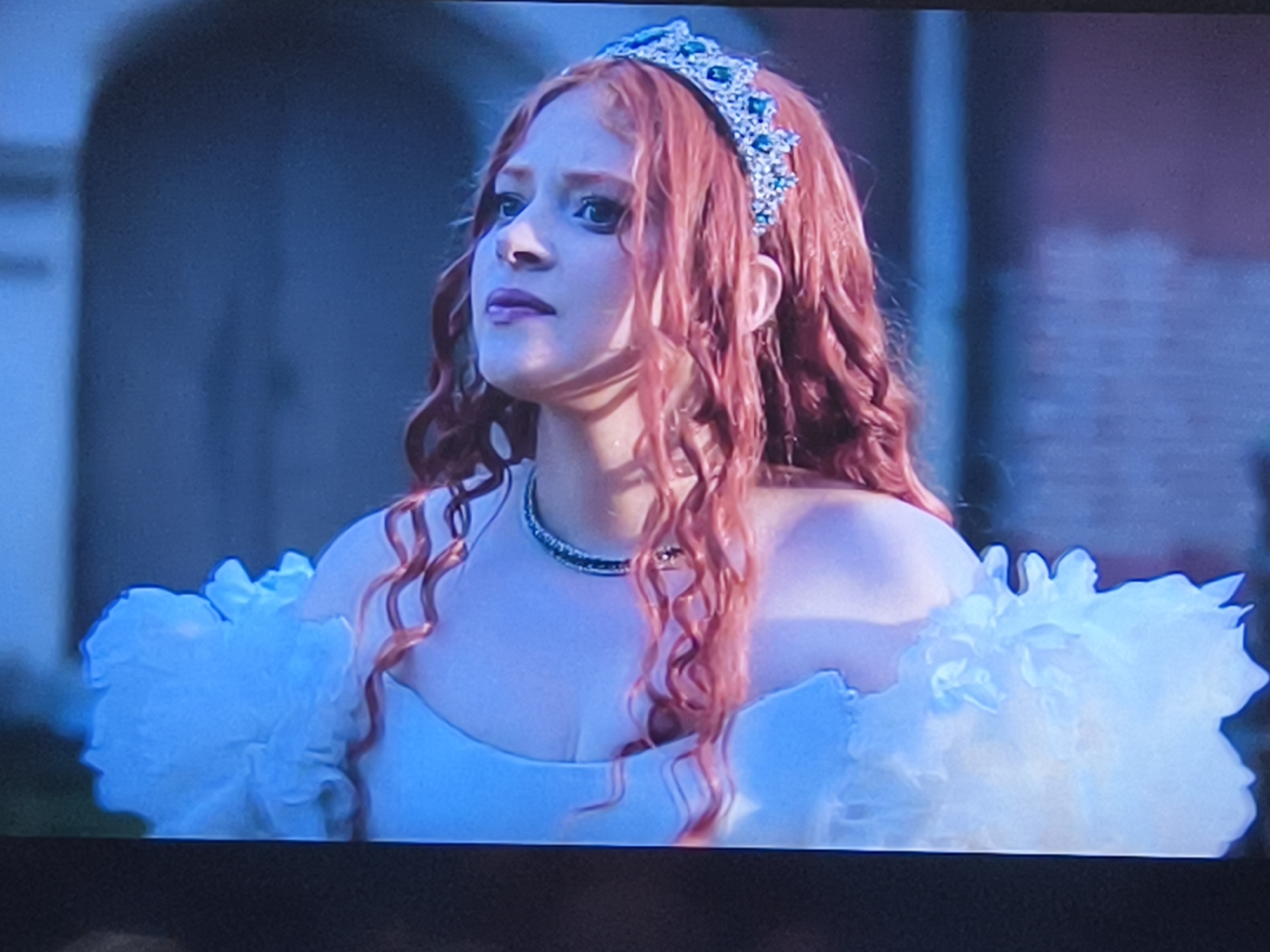A medium shot captures a woman with red, wavy hair adorned with a jeweled tiara. She wears a white dress with large, fluffy white shoulders. The woman's face shows a contemplative expression as she looks up and to the left. A necklace is visible around her neck. Behind her, an indistinct structure suggests a building. The environment appears muted with a blue tone. There is no verifiable information about Petah Tikva, Israel in the image.