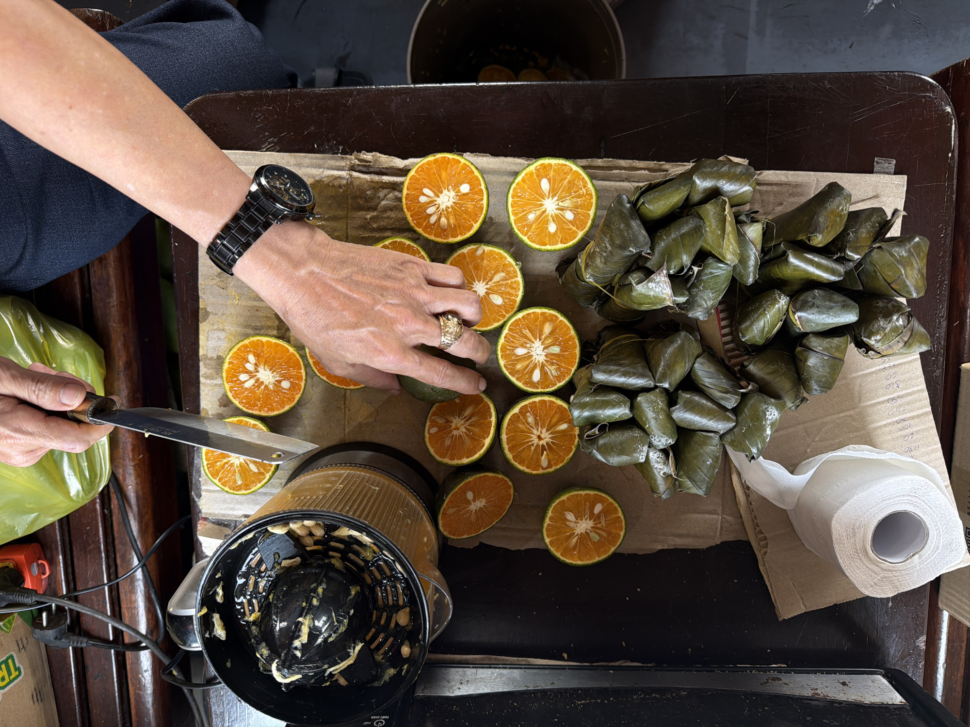 The image, taken from a high-angle perspective, shows a person preparing citrus fruit, possibly oranges, in what appears to be a domestic setting, potentially in the city of Huế, Vietnam. The person, wearing a black watch and a gold ring, is using a knife to cut the oranges in half on a wooden surface, likely a cutting board or table. Several orange halves are already prepared and arranged on the board. A black citrus juicer is positioned near the sliced fruit, suggesting the oranges are being prepared for juicing. Adjacent to the oranges, a stack of leafy green packages, possibly wrapped food items, are present. A roll of white paper towels rests near the food packages. The background shows parts of furniture and possible food packaging, providing further context to the domestic setting.