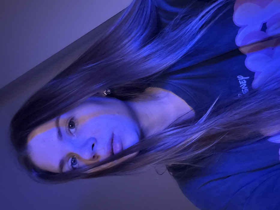 A young woman with long, light brown hair is angled in the frame. She wears a dark shirt with "Disney" written on it. A white, cloud-like object is partially visible. The image is bathed in blue light, highlighting her fair skin and features. She has blue eyes and a neutral expression. An earring is visible in her left ear.