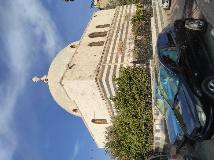 The image depicts a street scene in Al-Qanawat Municipality, Syria, dominated by a large stone building under a partially cloudy blue sky. The central structure features a prominent, light-colored dome topped with a decorative finial. Its facade is constructed from light-toned stone blocks, with horizontal sections of striped ablaq masonry visible on the lower levels. Multiple arched windows with light-colored frames are present on the building's upper walls.
In the foreground, a paved street runs horizontally, with several vehicles parked along it. A black two-door hatchback, resembling a Peugeot 206, is prominently positioned in the lower-center, facing right. Behind it and to the left, a light blue sedan is parked. The rear portion of a silver or grey vehicle is partially visible on the far left. To the far right, a white sedan is partially visible further down the street.
Separating the street and the main building is a low wall or planter, partially adorned with affixed papers or posters, topped by a dark metal fence. Behind this fence and wall, dense green foliage, consistent with bushes or small trees, lines the base of the building. No individuals are visible in the scene.