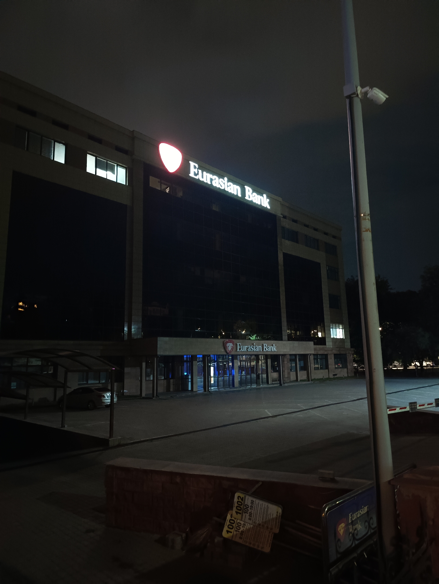 This image shows the Eurasian Bank building in Almaty, Kazakhstan, at night. The building is illuminated, with its name clearly visible in large, bright letters on the facade. The architecture is modern, featuring large glass windows and a light-colored stone exterior. In the foreground, a parking lot is visible, with a few cars parked and what appears to be a security barrier. A lamppost with a security camera attached is also present. The sky is dark, suggesting it is nighttime. The scene is quiet and still, with no people visible. The overall impression is one of a secure and well-maintained financial institution.