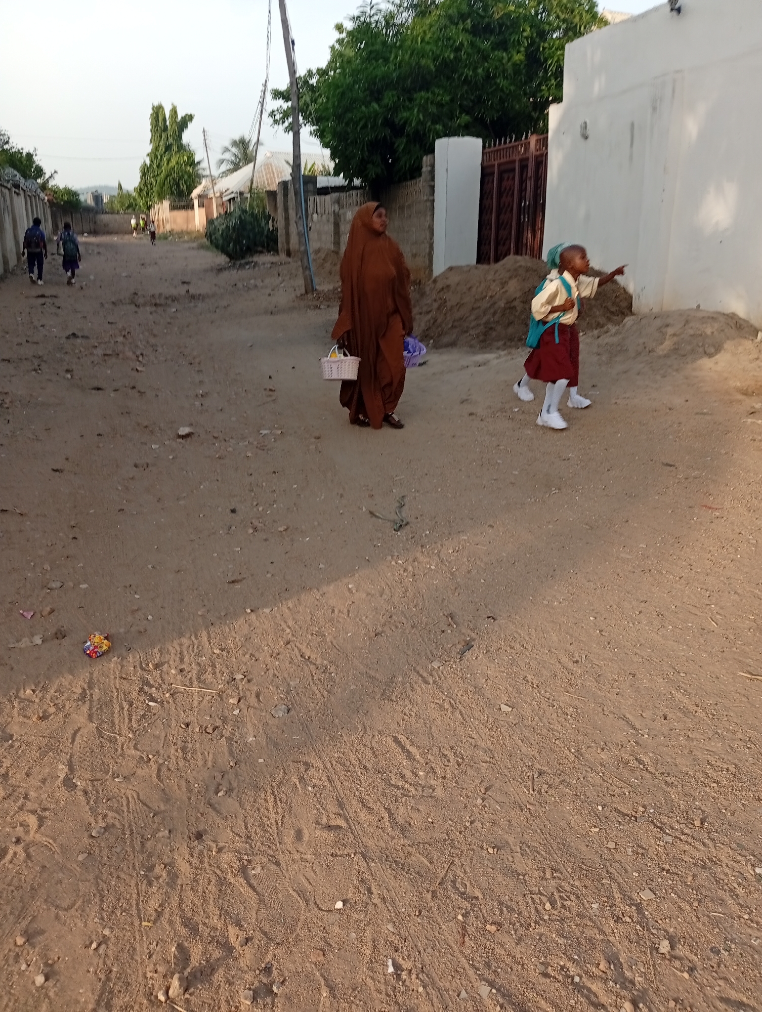 Location: Adankolo, Nigeria. A dirt road runs through the scene. A woman in a brown hijab carries a white basket. A child in a school uniform with a turquoise backpack points. Other people walk in the distance. A wrapper lies on the ground. Buildings and trees line the road. Sand piles are near a white wall.