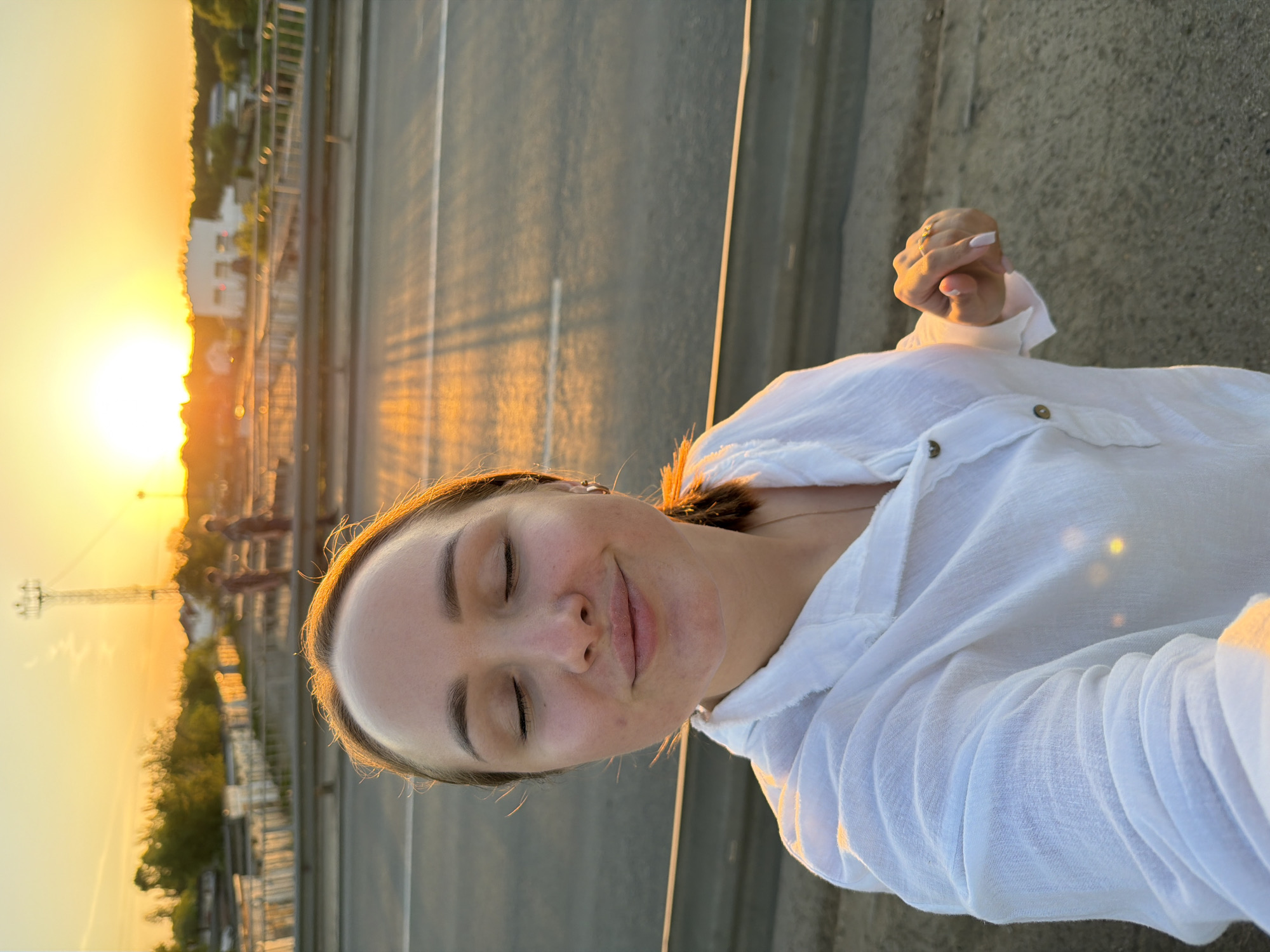 A selfie taken in Chaikovsky, Russia, at sunset. A woman with closed eyes is smiling at the camera. She is wearing a white, collared shirt with rolled up sleeves. The background shows the bridge over the Kama River and the setting sun. The lighting casts long shadows on the bridge and a golden hue on the woman's face.