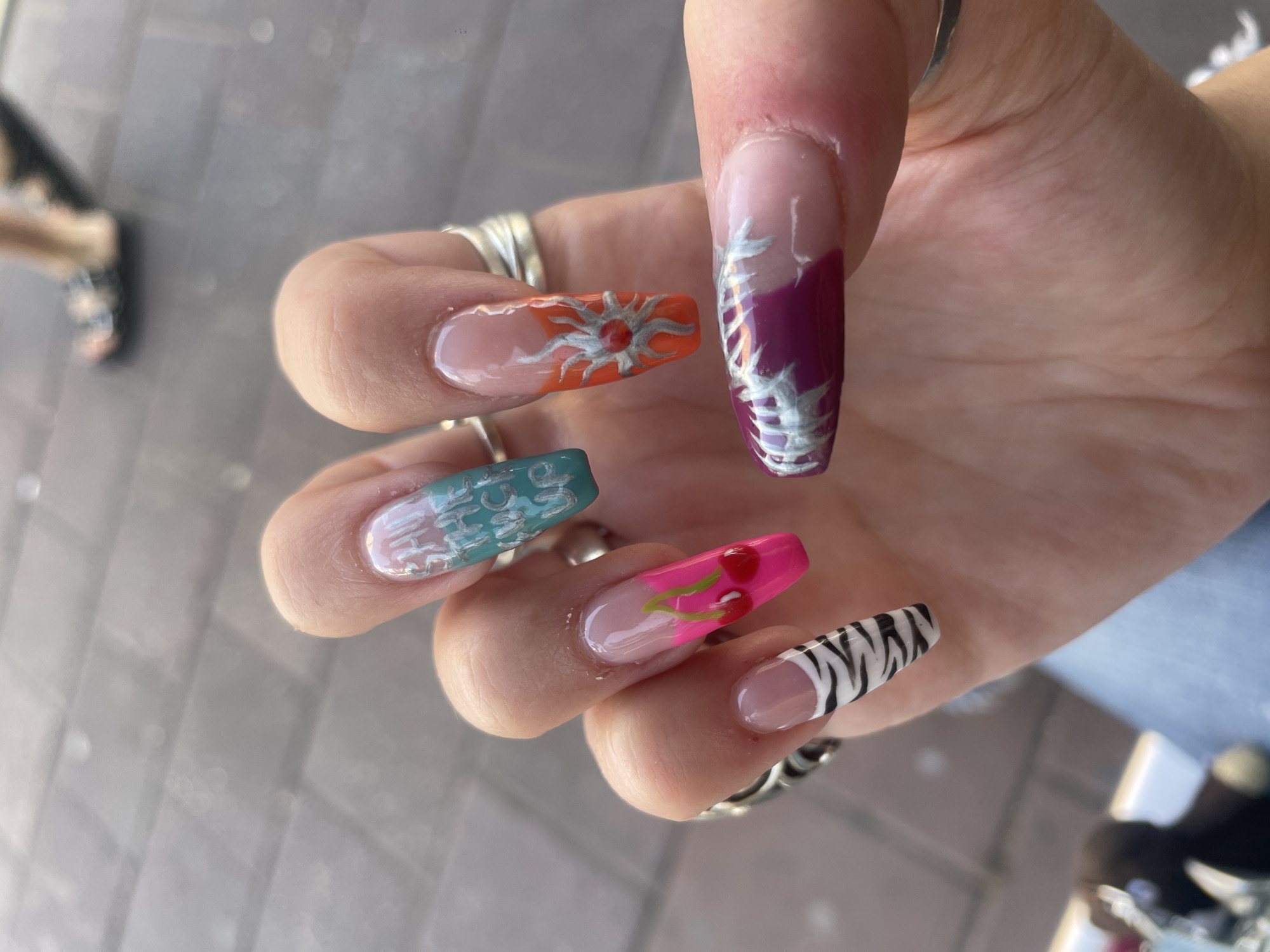 The image displays a close-up of a person's hand with elaborately decorated fingernails. The background is blurred, showing a pavement surface and the legs of another person in sandals. The nails feature a variety of designs: one has a zebra print, another depicts bright pink cherries with green stems, and a third is adorned with an orange sun-like motif. One nail has white lettering on a gradient of teal and pink, appearing to read "SAVE THE FUCK UP". Another nail has a design in purple and pink with silver accents. The hand is adorned with multiple silver rings. The setting appears to be outdoors, possibly on a street or public walkway. No specific location in Ramat Gan, Israel is discernible from the image alone.