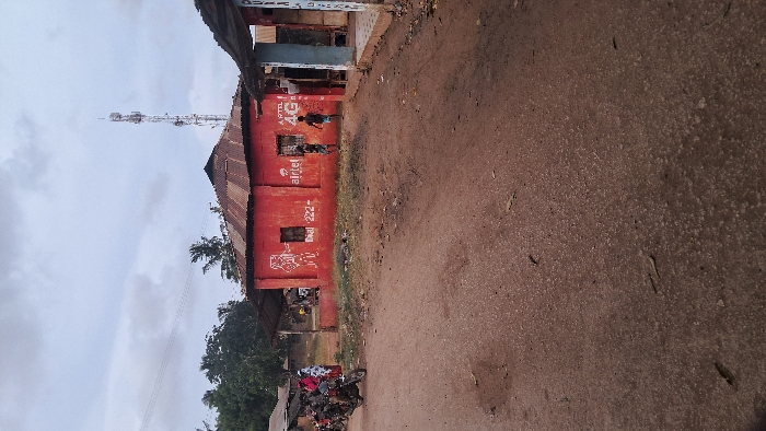 The image captures a rural or semi-urban street scene under an overcast sky. The primary focus is a single-story building on the left, painted a distinctive red or reddish-brown, with a weathered, corrugated metal roof that appears dark brown. The building's walls feature white painted text and logos. Prominently displayed are "APTL" and "4G," followed by the "airtel" logo. Further along the wall, the text "Dial *222#" is visible, along with a graphic resembling a mobile phone or network signal icon.
Several individuals are present near the red building. Two figures, possibly children or young adults, are standing or moving close to the "APTL 4G" and "airtel" branding. Another person is partially visible near what appears to be an entrance or stall further left along the building.
In the foreground, a reddish-brown dirt or gravel road stretches across the lower and right parts of the frame. The road appears unpaved, showing texture and small scattered debris. On the far left, a motorcycle is parked or stopped, its seat covered with a distinctive patterned fabric, possibly red and white.
Behind the red building and extending towards the left side of the image, various green trees and foliage are visible, including what appears to be a palm tree. A tall, metallic communication tower, equipped with antennas and dishes, stands prominently in the background against the cloudy, grey sky, signifying modern telecommunication infrastructure in the area. The sky is uniformly grey, indicating an overcast day without direct sunlight.