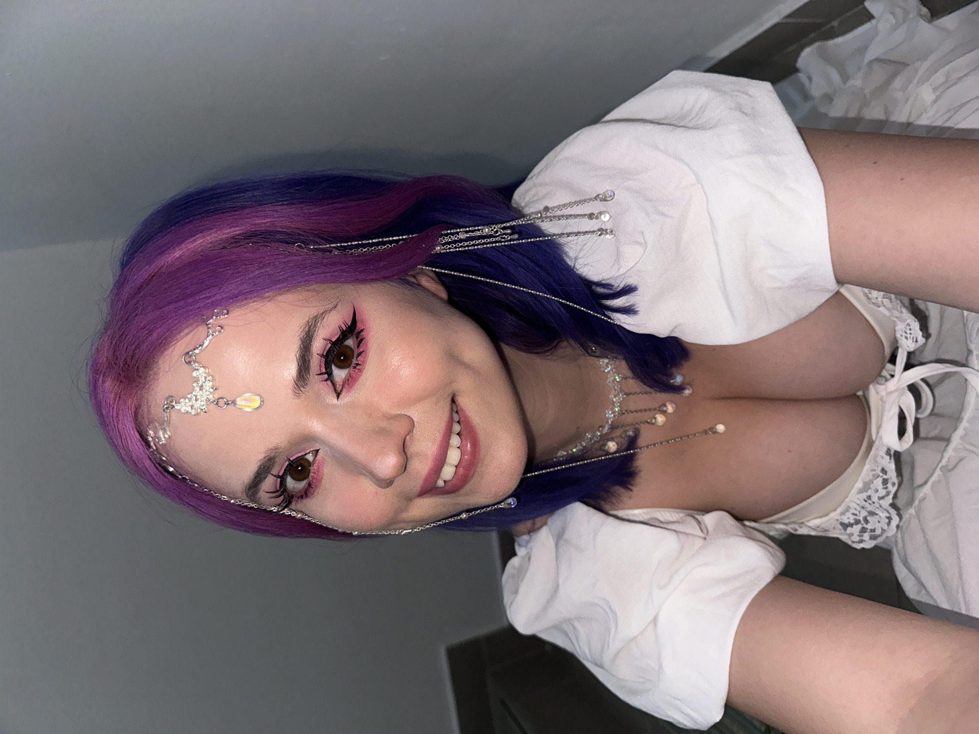 The image shows a woman with purple and blue hair smiling and looking into the camera. She has dramatic pink eyeliner and a decorative silver headpiece. She is wearing a white peasant-style top with a ruffled neckline. The background consists of a gray wall and a small section of what appears to be a floor. No details suggest the scene is located in the City of Zagreb, Croatia.