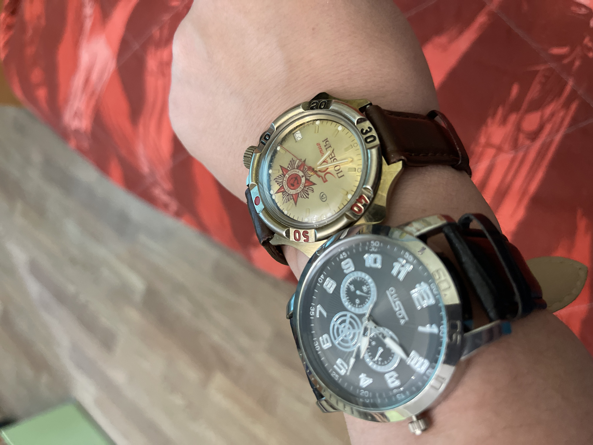 The image shows a person's arm with two wristwatches on it. One watch has a brown leather strap and a gold-colored face featuring Soviet-era imagery including a red star and the inscription "ПОБЕДЫ" (Victory), a reference to the Great Patriotic War. The other watch has a black face with multiple sub-dials, a silver-colored case, and a black leather strap. In the background, there is a textured surface in shades of red and tan that might be a bedspread. There is also a floor with light wood paneling. No information is available regarding the location in the city of Moscow, Russia.