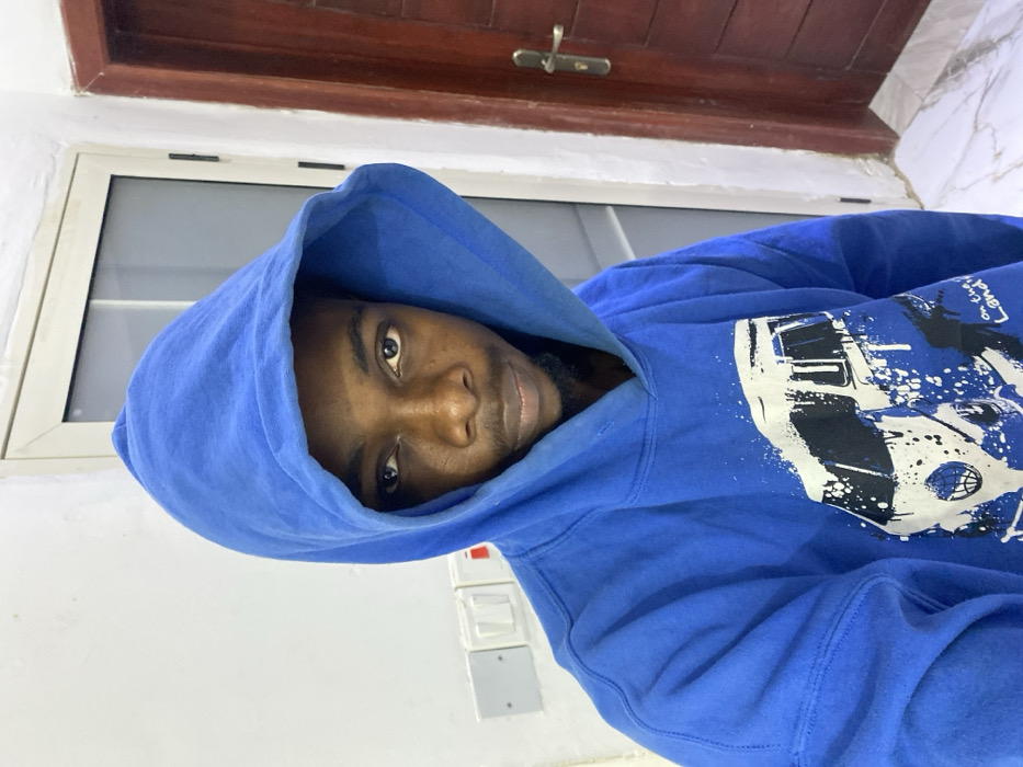 The image features a dark-skinned man wearing a blue hooded sweatshirt (hoodie), looking directly into the camera. He has a beard or stubble, and the hood is up, framing his face. The blue hoodie has a prominent graphic design on the right chest and shoulder area, rendered in white and black. This design incorporates elements resembling the front of a vehicle (possibly a truck or car), a basketball, and paint splatters. Above these, the text "On The LAND" is clearly visible.
The background consists of a plain white wall. To the man's left, a white-framed window is present, allowing natural light into the space. Above the window, there is a dark wooden structure, possibly a door frame or an overhead beam, with a metallic latch visible. To the man's right, on the white wall, a set of white and gray electrical switches or outlets can be seen. The setting appears to be an indoor environment. The man maintains a calm, neutral expression. The image is oriented 90 degrees clockwise from what would be a standard upright portrait. The location context is Joma, Ghana.