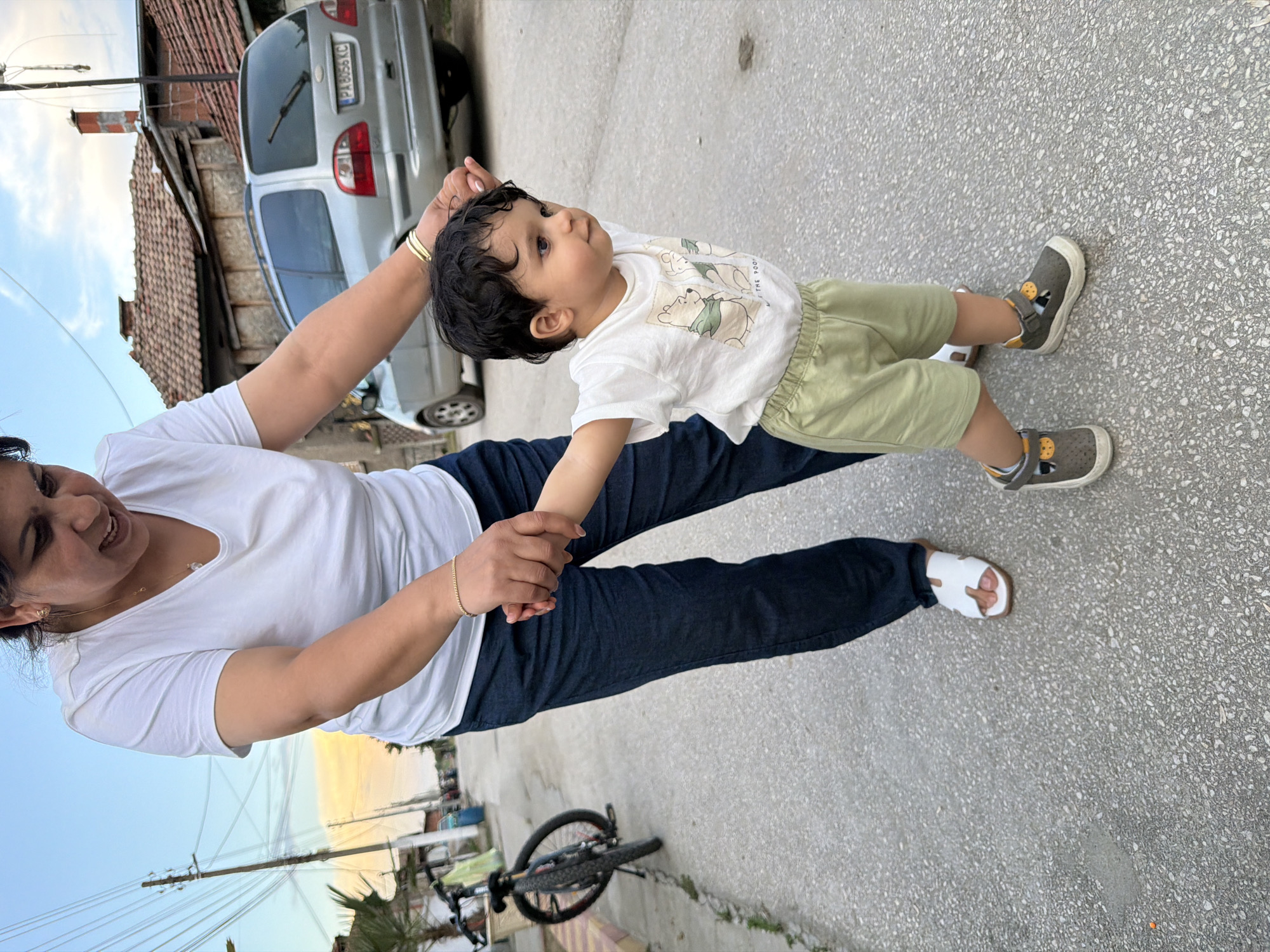 An outdoor photograph, rotated ninety degrees counter-clockwise, depicts an adult woman assisting a young child in walking on an asphalt or concrete surface. The woman, with dark hair, wears a white V-neck t-shirt, dark blue jeans, and white sandals. A gold bracelet adorns her right wrist. She is smiling as she holds the child's hands.
The child, with dark curly hair and blue eyes, is dressed in a white t-shirt featuring a Winnie the Pooh design (Pooh Bear and Piglet, with the text "THE POOH" visible), light green shorts, and grey and yellow shoes. The child looks upwards and slightly to the right, showing an expression of curiosity or attention.
The setting appears to be a residential street or alley in Sinitovo, Bulgaria. In the background, an older building with a red tiled roof and a chimney is visible. A silver minivan-style car, possibly a Ford Galaxy, is parked, and its Bulgarian license plate, "PA 8056 KC," confirms the region as Pazardzhik province. Utility poles and overhead wires crisscross the sky. A bicycle lies on its side on the ground in the lower right foreground. The sky displays a mix of blue and softer, warmer hues, suggesting the time of day is late afternoon or early evening under clear weather conditions.