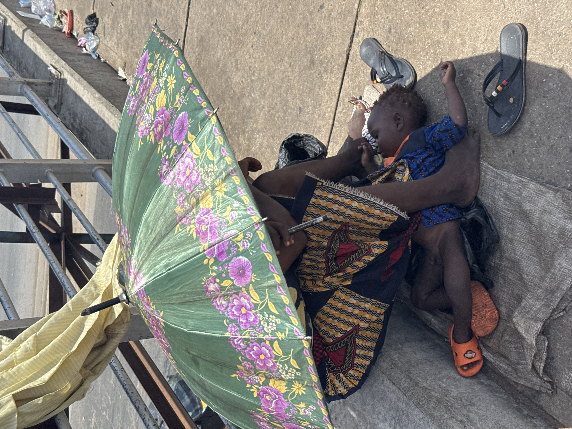 The image presents an overhead view of an adult and a young child resting outdoors, likely on a concrete surface in Onitsha, Nigeria. Bright sunlight and strong shadows indicate it is daytime, with a prominent green umbrella, adorned with purple and yellow floral patterns, open and providing shade over the figures.
An adult, mostly obscured but identifiable by an arm embracing the child and a patterned garment featuring orange, black, and white geometric designs, is visible. Partially readable text on this fabric includes "APPIES" and "BLOCK". The adult holds a black pen in one hand. The child, appearing to be a toddler with short dark hair, lies across the adult's lap, seemingly peaceful or asleep, wearing a blue patterned top and orange sandals that have a small white cartoonish design.
Adjacent to the child's head on the concrete are two dark-colored flip-flops, one featuring multicolored stripes on its strap. The individuals are positioned on a grey, textured fabric, possibly a tarp or blanket. The background to the upper left reveals a concrete ledge or step littered with various pieces of plastic trash. Metal railings, possibly from an overpass or structure, and a draped yellow fabric are also visible on the left side of the frame. The scene depicts a moment of rest or seeking shelter from the sun in an urban outdoor environment.