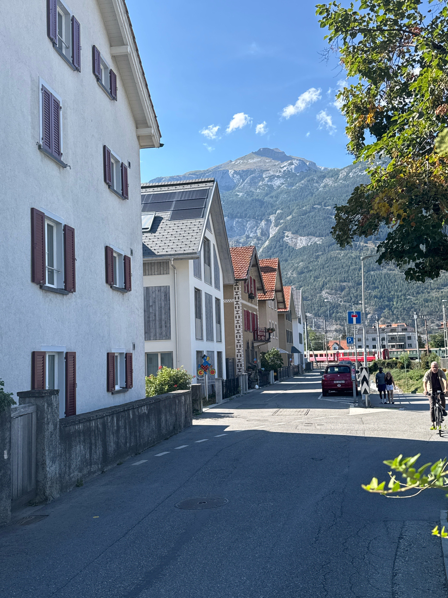 The image depicts a street scene in a town nestled at the base of mountains. In the foreground, a white house with dark red shutters lines the left side of the street. Further down the street, a row of multi-story buildings with red-tiled roofs is visible. A red car is parked on the right side of the street. In the background, a train with a red exterior is traveling on tracks. People are present in the scene, with a man on a bicycle on the right side of the street and a couple walking on the sidewalk. The sky is clear and blue with a few scattered clouds. The overall environment is characterized by residential buildings, infrastructure like train tracks and roads, and a mountainous backdrop.
