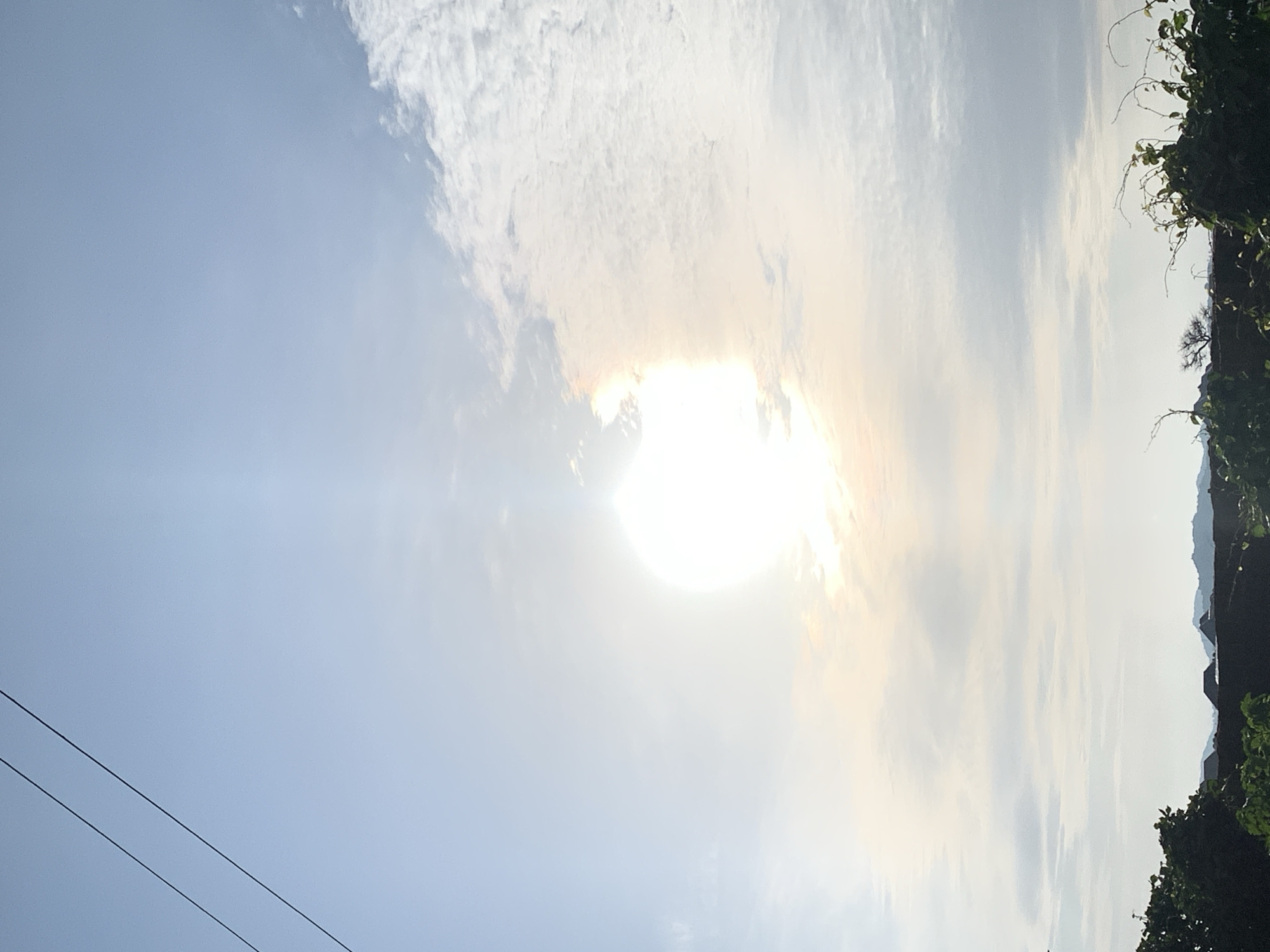 A bright, hazy sun is visible in the sky above a landscape in Kuru Karama, Nigeria. The sun is partially obscured by clouds, creating a halo effect. Two power lines stretch across the upper left portion of the image. The lower part of the image shows a dark silhouette of trees, bushes, and buildings against the brighter sky. Distant hills are visible on the horizon. The overall impression is of a bright, possibly overcast day.