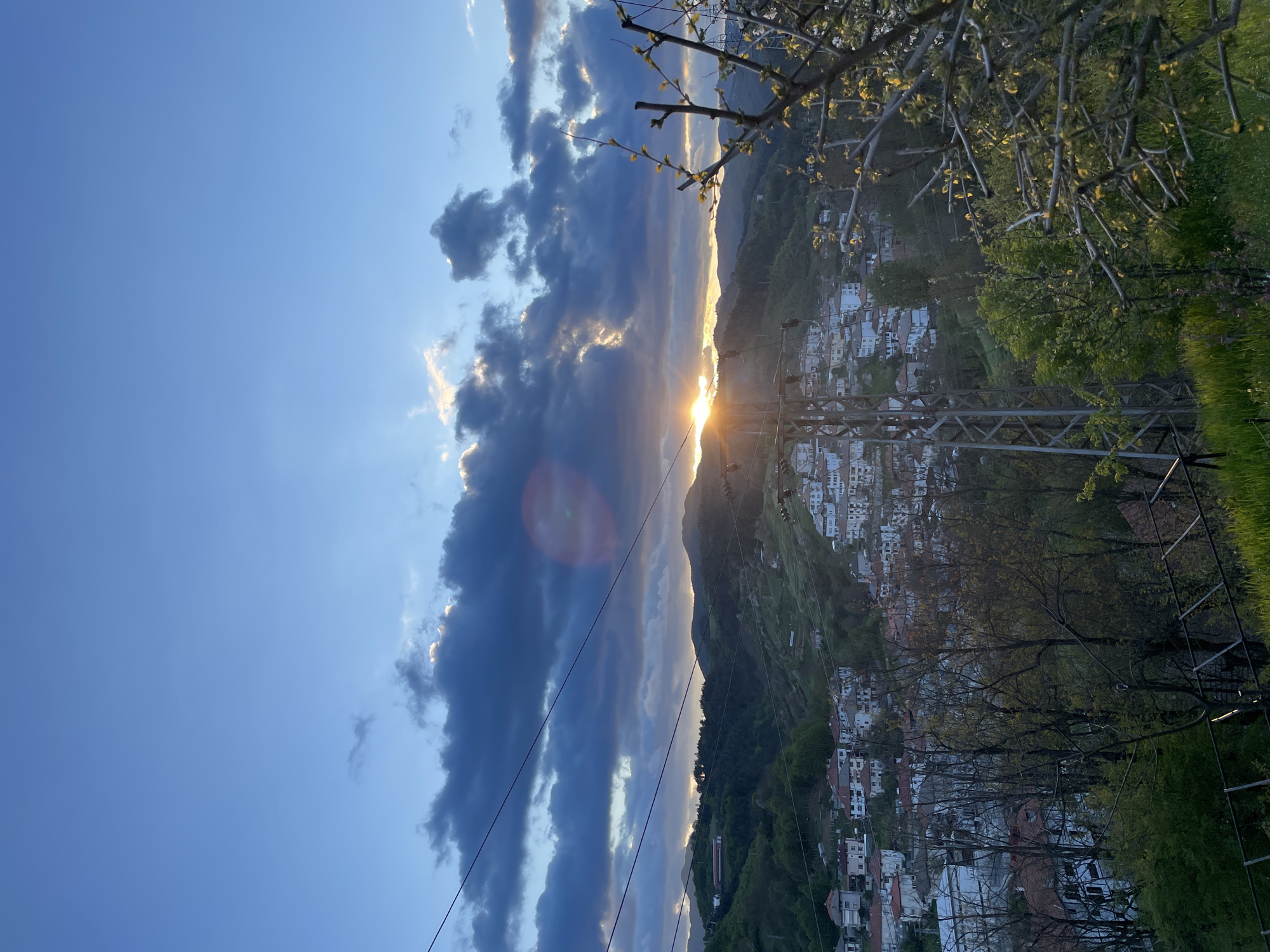 The image shows a sunset view in the city of Zlatograd, Bulgaria. Dark clouds with sun rays shining through them are visible in the sky. Below, there is a mountainous landscape with a town built on the slopes, with residential buildings with red roofs. Trees and power lines are in the foreground, and the sky is clear blue in some areas.