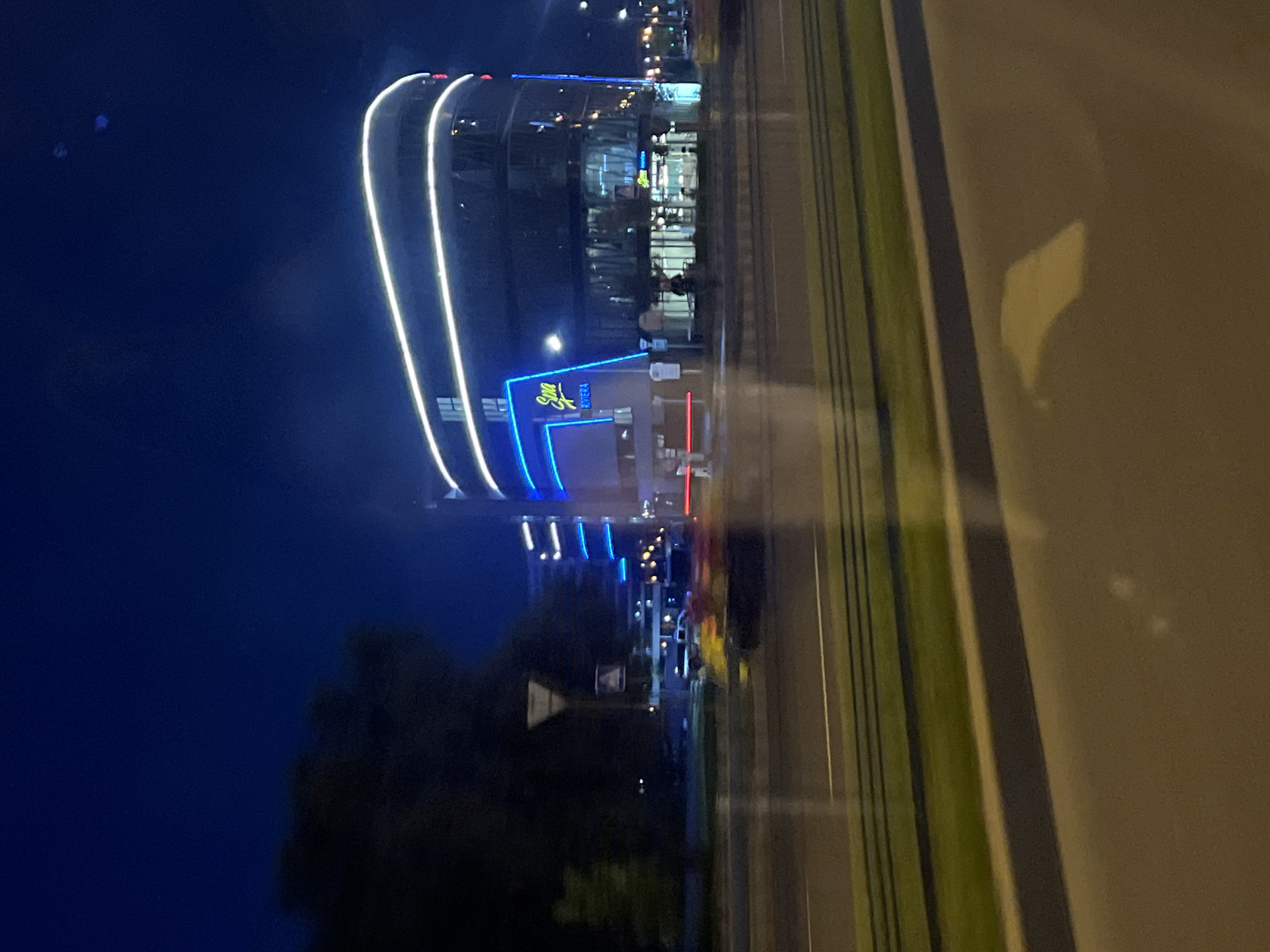 The image depicts the "Zvezda" Riviera shopping center in Minsk, Belarus, at night. The building is characterized by its modern, curved architecture and prominent blue and white neon lighting. Several lanes of traffic are visible in the foreground, with blurred car lights indicating movement. Green grass borders the road, and a dark silhouette of trees frames the left side of the scene. The overall impression is that of a vibrant urban environment at night, with the shopping center serving as a focal point illuminated by artificial lights.