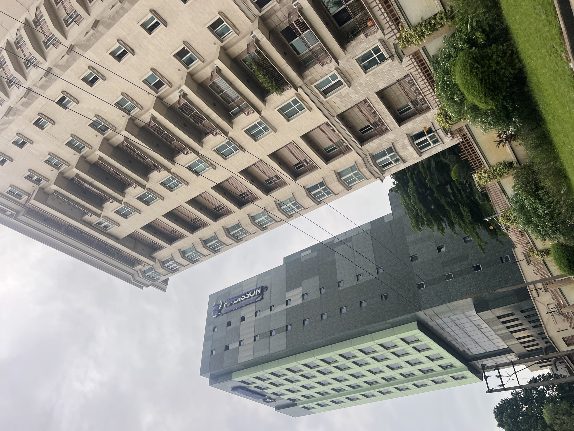 In the city of Ikoyi, Nigeria, this image captures a view of two modern buildings against an overcast sky. The building on the left is a tall, slender structure with a facade of alternating light green and dark gray panels. Several small, dark square windows are visible on the dark gray sections, while a grid of rectangular windows covers the green sections. A prominent banner with the word "RADISSON" in white lettering on a dark blue background is affixed vertically to the side of this building. The building on the right is a beige, multi-story residential or office building with a repeating pattern of balconies and windows. Each balcony has a brown metal railing. The lower part of the image shows a well-maintained green lawn and various shrubs and trees. A low, ornate brown fence with climbing plants runs along the edge of the property. Electrical poles and wires are also visible in the foreground, indicating urban infrastructure. The perspective is low-angle, emphasizing the height of the buildings.