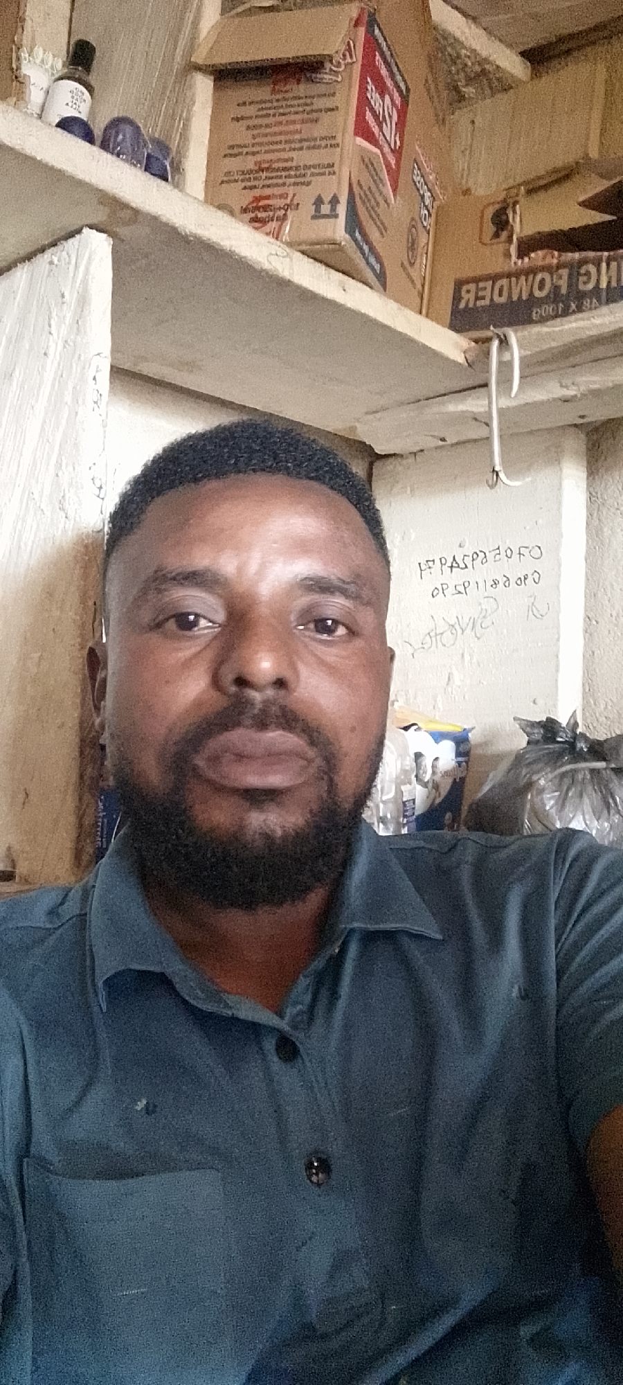 The image shows a close-up, selfie-style shot of a dark-skinned man with short, black hair and a full beard. He is wearing a dark blue collared shirt with two black buttons visible. The background appears to be a storage area with shelves stacked with various items, including cardboard boxes, bottles, and plastic bags. One box is labeled "Baking Powder." In the upper right corner of the image, there is handwritten text on a white wall. The man's facial expression is neutral. The lighting suggests an indoor environment, possibly a shop or storeroom. There is no discernible indication of the specific location within Durumi, Nigeria.