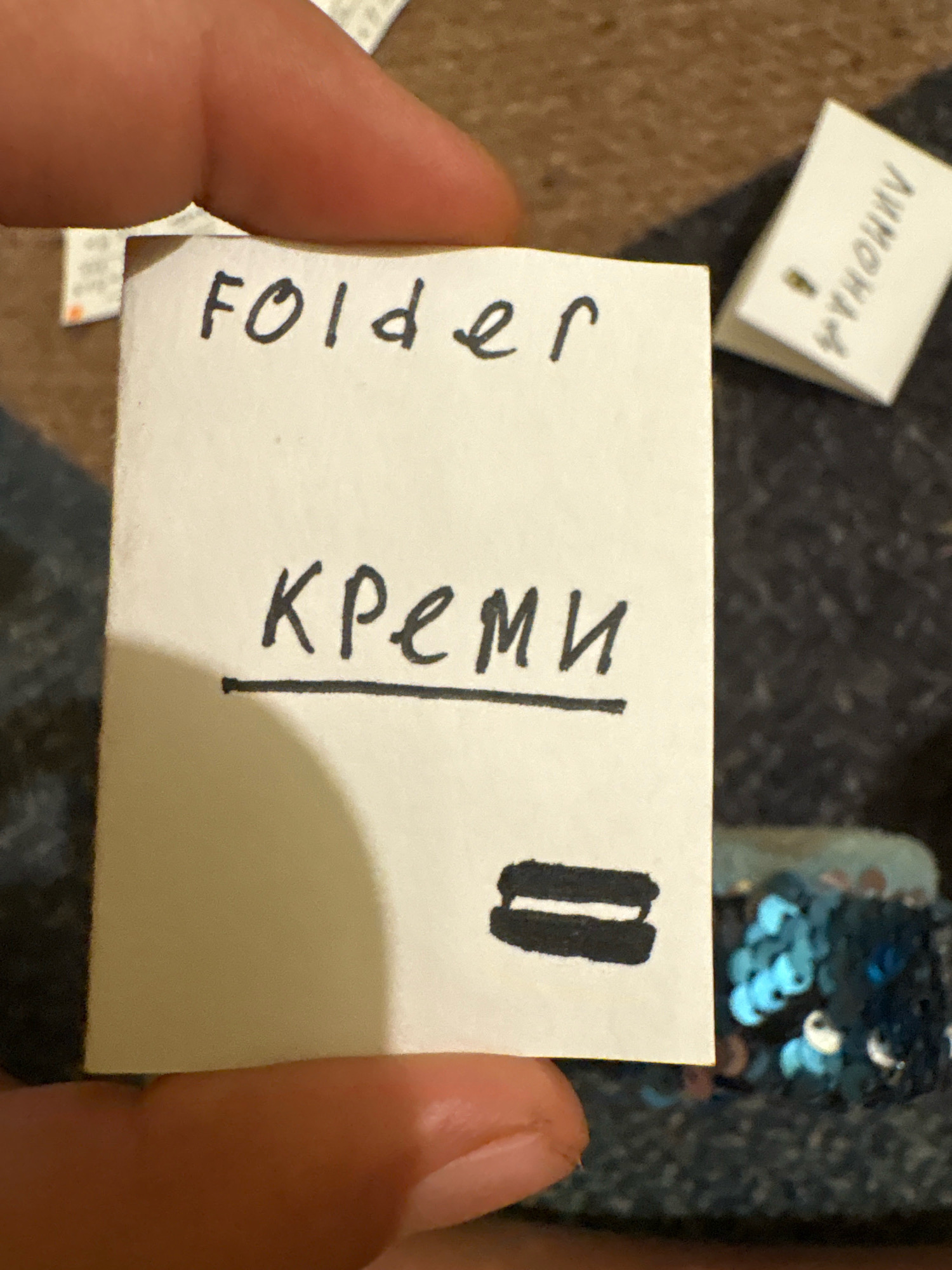 The image is a close-up shot of a hand holding a small, white piece of paper with handwritten text. The text on the paper is in English and Russian. The top line of text reads "folder" in English. Below that, it reads "креми" in Russian, with a line underneath. At the bottom of the paper, there is a symbol that resembles two horizontal lines stacked on top of each other.
In the background, there is a small, folded white label with Cyrillic text that appears to read "Лимона" (Limona). To the right of the paper, there is a blurred area with dark fabric that has scattered blue and silver sequins. The scene is likely indoors, possibly on a table or flat surface, given the texture of the background.
The image does not contain any information that would place it in Ashvem, India, or any specific city.