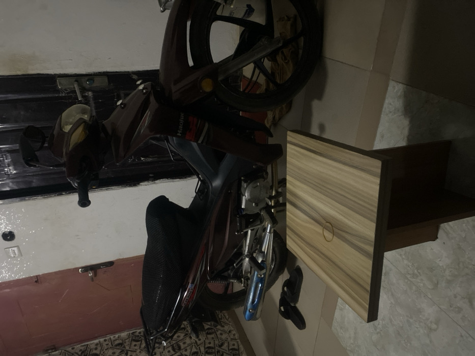 A maroon scooter stands beside a wooden cabinet with a door. The scooter features a black seat, a headlight with a gold tint, and a blue exhaust pipe. One wheel is visible, and what appear to be sandals are situated near the scooter's base, close to the cabinet. The background includes a tiled floor, a door, and parts of a wall. The image is rotated counterclockwise.
