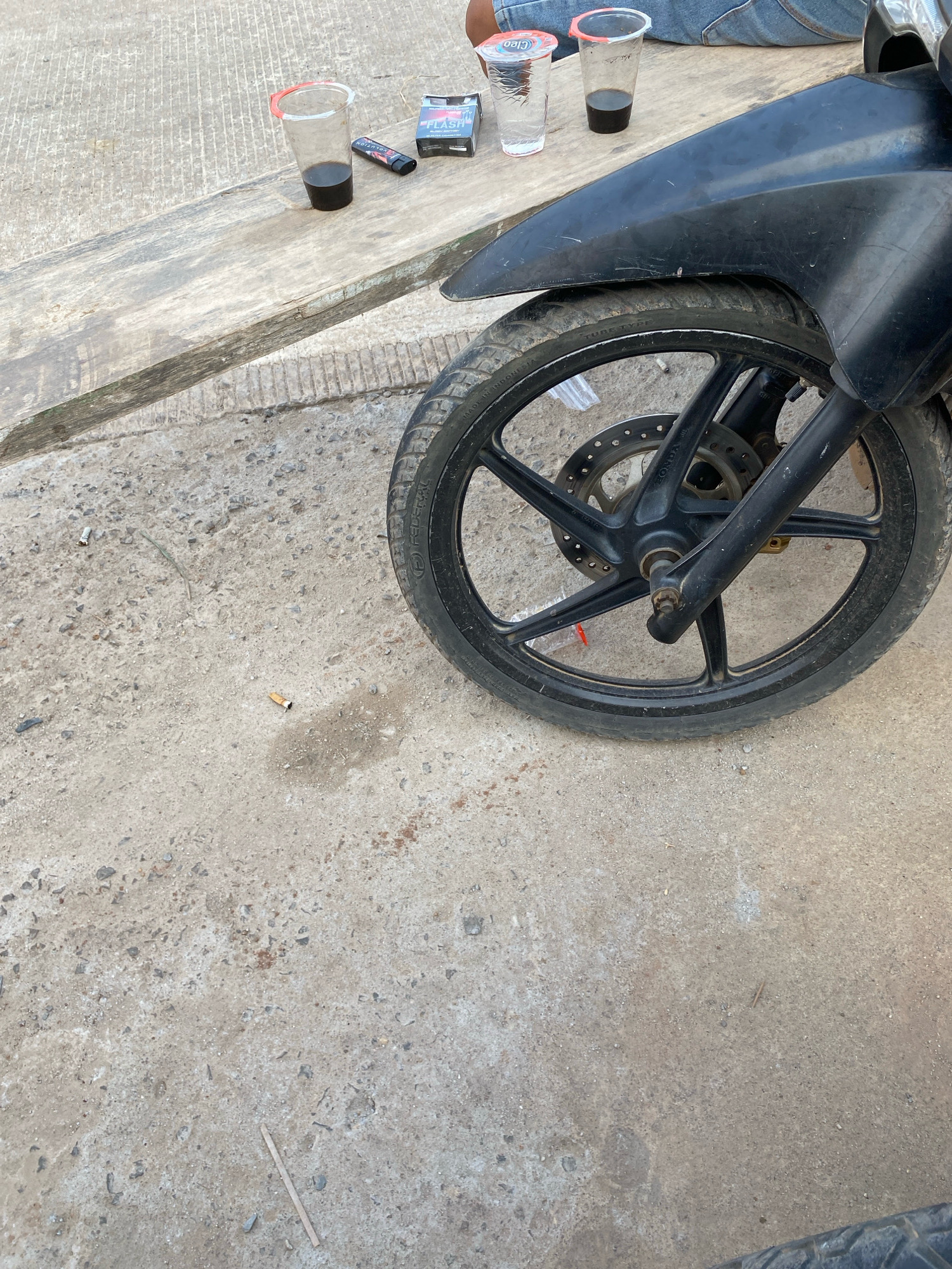 A close-up, low-angle shot captures a motorcycle's front wheel and fender on a concrete surface. To the left, a wooden plank rests on the ground. On the plank, a cigarette pack labeled "FLASH," a lighter, and two clear plastic cups containing dark liquid and clear liquid are visible. The ground surrounding the plank and motorcycle is concrete, with scattered cigarette butts and small debris. A partial view of blue denim pants suggests someone is sitting nearby, possibly near the plank and drinks. The overall environment appears to be an outdoor urban setting.