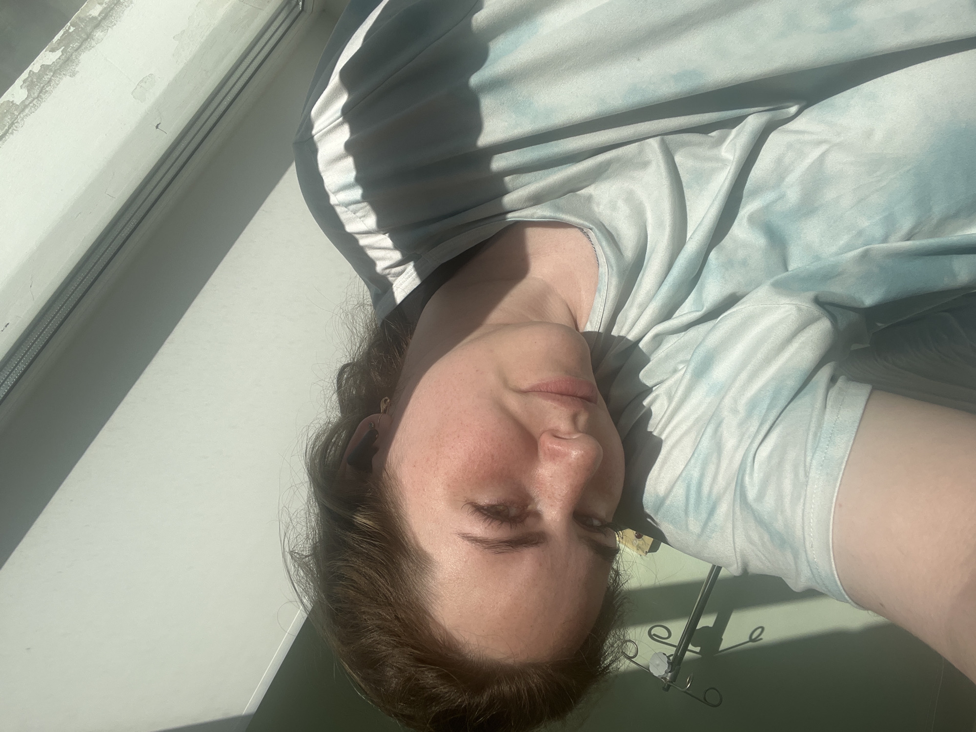 A woman with reddish-brown hair is lying on a white surface, possibly a bed, near a window. She is wearing a light blue and white tie-dye shirt. Her face is visible, and she appears to be indoors. An IV pole is partially visible in the lower right corner. The setting appears to be a hospital room or healthcare facility. The location in the city of Chaikovsky, Russia, is not discernible from the image.
