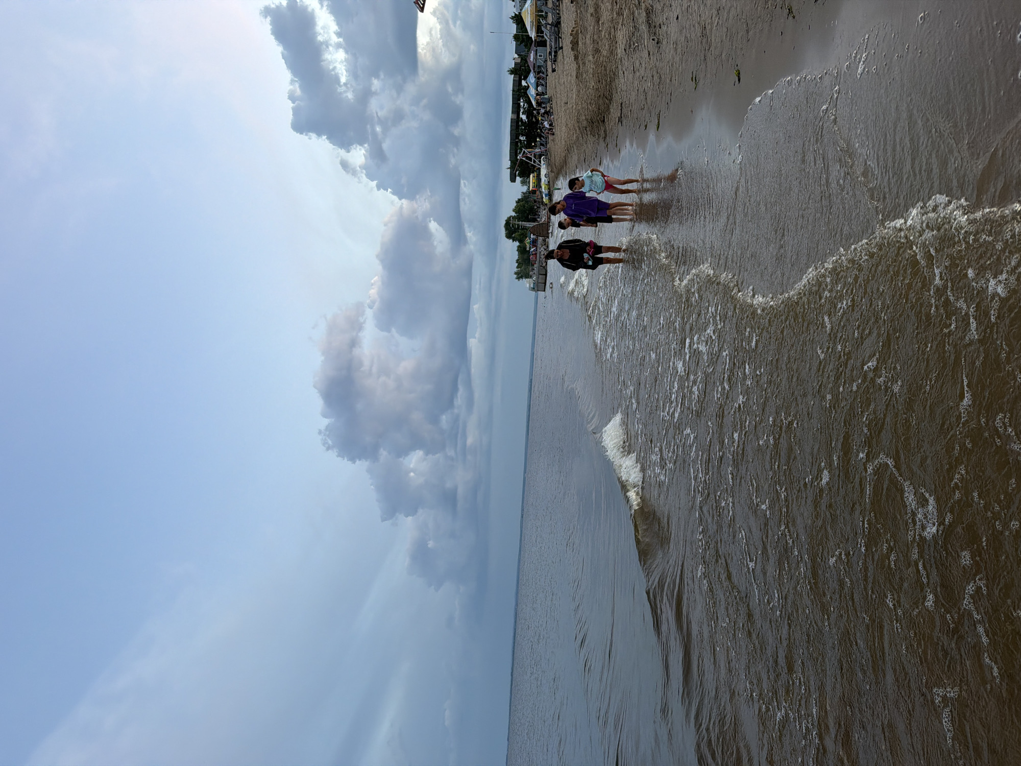 A vertical, eye-level, full shot captures a group of four people walking along the shoreline of the Volga River in Laishevo, Russia. The water laps at their ankles as they walk to the right of the frame. The beach is sandy, with a few scattered pieces of seaweed. In the distance, there are several tents and buildings, likely part of the beach resort. The sky above is a mix of blue and gray clouds, suggesting a partly cloudy day. The lighting is soft and diffused, indicating it is either early morning or late afternoon. The overall mood of the image is serene and peaceful.