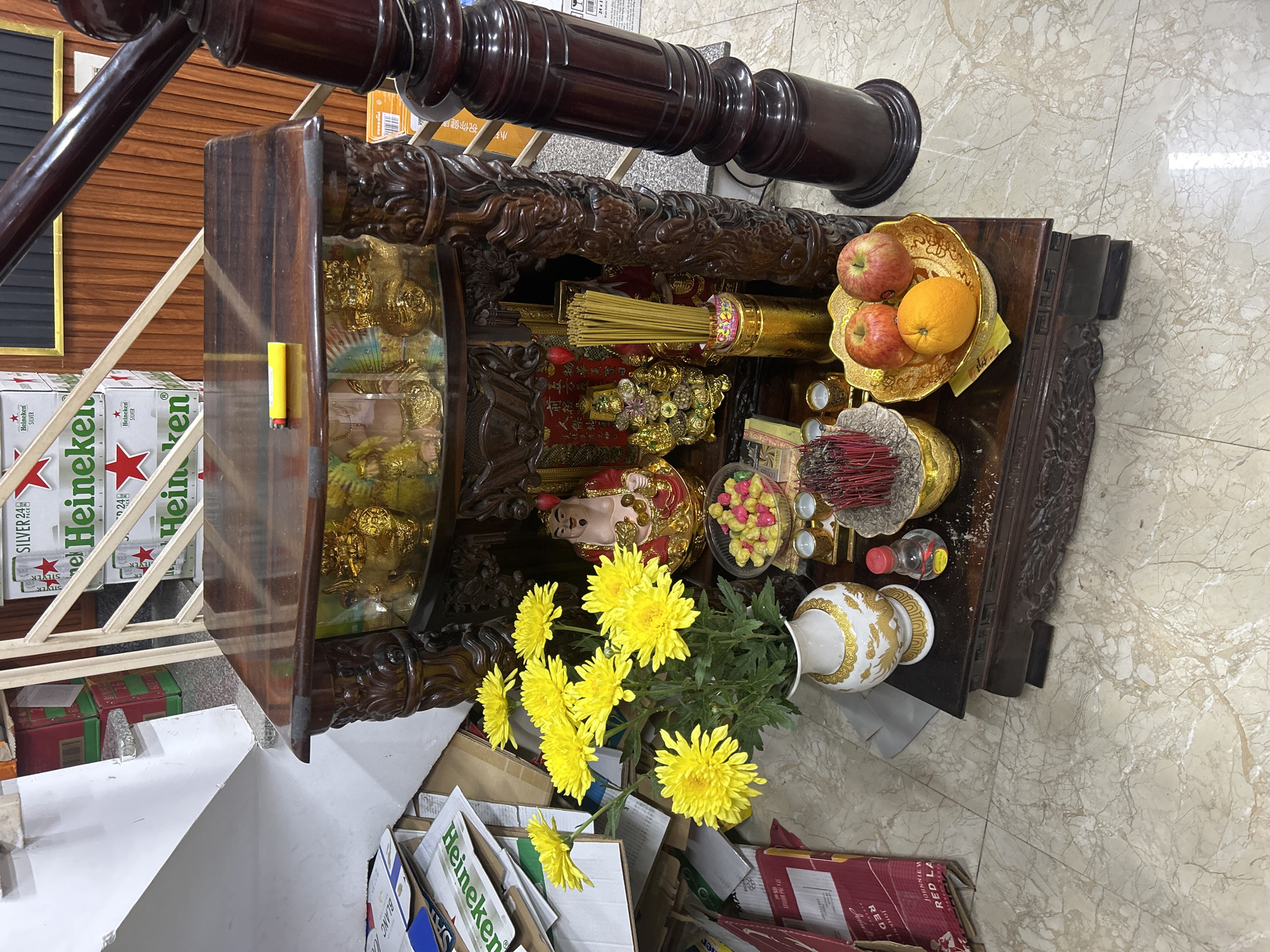 This media file, captured in Nha Trang Ward, Vietnam, depicts an indoor scene centered around a richly carved dark wooden altar or shrine. The altar is adorned with various offerings, suggesting a religious or spiritual practice. Golden statues, including what appears to be a Laughing Buddha, are prominently displayed. Bundles of incense sticks are placed in golden holders, alongside a golden bowl containing red apples and an orange. A white vase holds a vibrant bouquet of yellow chrysanthemum flowers, and small cups likely intended for liquid offerings are also present. Colorful candies or sweets are arranged in a clear container, and a small bottle and a yellow lighter are also visible on the altar.
The background and foreground provide additional context. Stacks of Heineken Silver beer boxes are visible on the left, while other cardboard boxes, some bearing "Heineken" and "Johnnie Walker Red Label" text, are on the floor. A dark wooden banister or decorative pillar runs diagonally across the upper part of the image. The flooring consists of light-colored marble or tiles. Text visible through OCR includes "Heineken SILVER," "JOHNNIE W RED LA" (likely for Red Label), and the Chinese phrase "小祝你健康" (Wishing you good health), which appears on a box in the background. The overall arrangement suggests an act of veneration or an offering ritual, a common cultural practice in Vietnamese homes or businesses. The scene is well-lit, indicating an indoor setting.