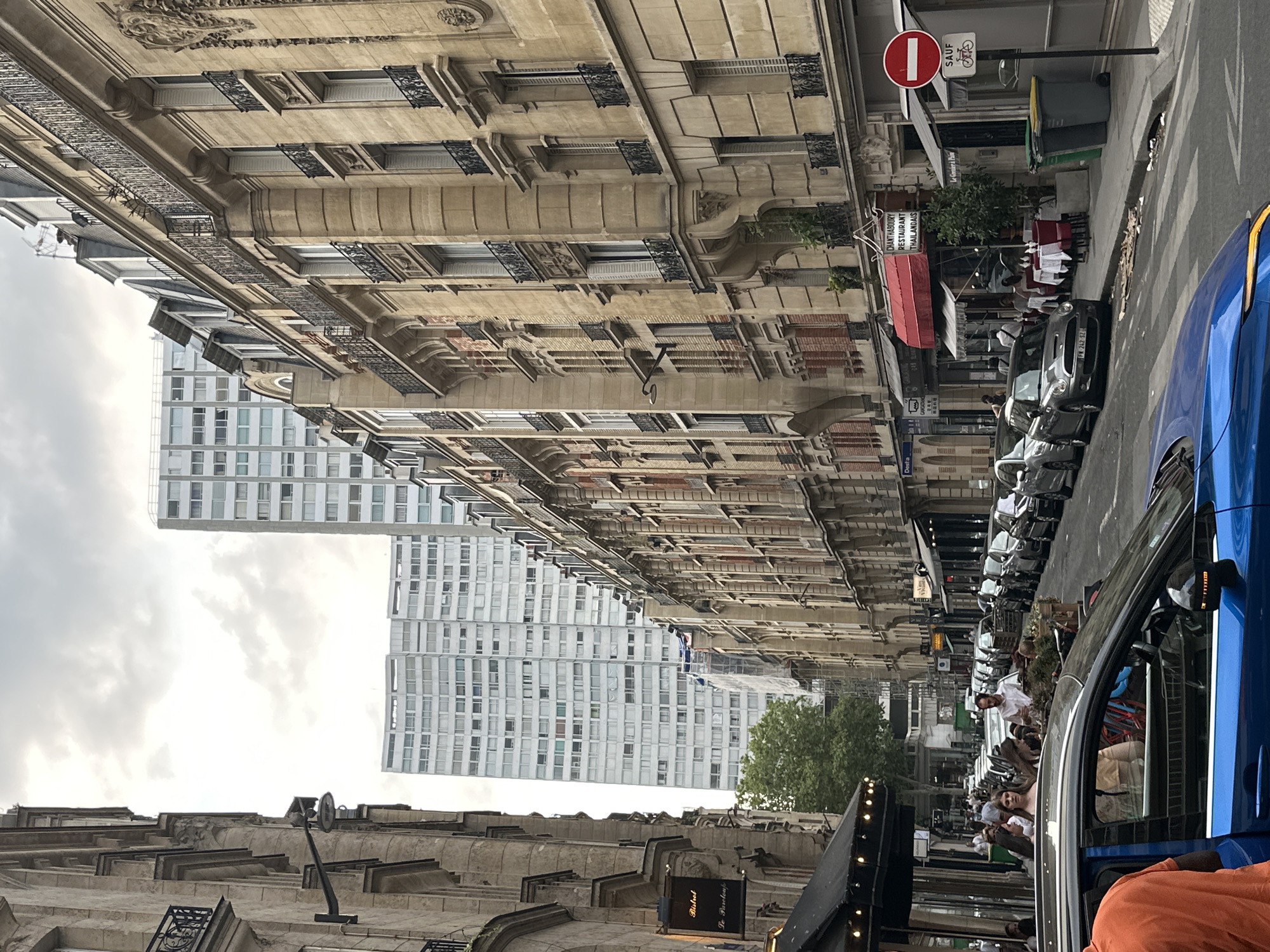 The image shows a street scene in Paris, France. The street is lined with tall, classic Parisian buildings with ornate facades and balconies. In the background, two modern high-rise buildings are visible. Cars are parked along the street, and a blue car is in the foreground, with a person in an orange shirt partially visible through the open door. Several people are seated at tables outside a restaurant with a red awning. A street sign indicates "CHANTHABOURY RESTAURANT THAILANDAIS" and a traffic sign shows a red circle with a white horizontal bar, meaning "no entry," along with a bicycle symbol below, indicating that bicycles are exempt. The sky is overcast with clouds.