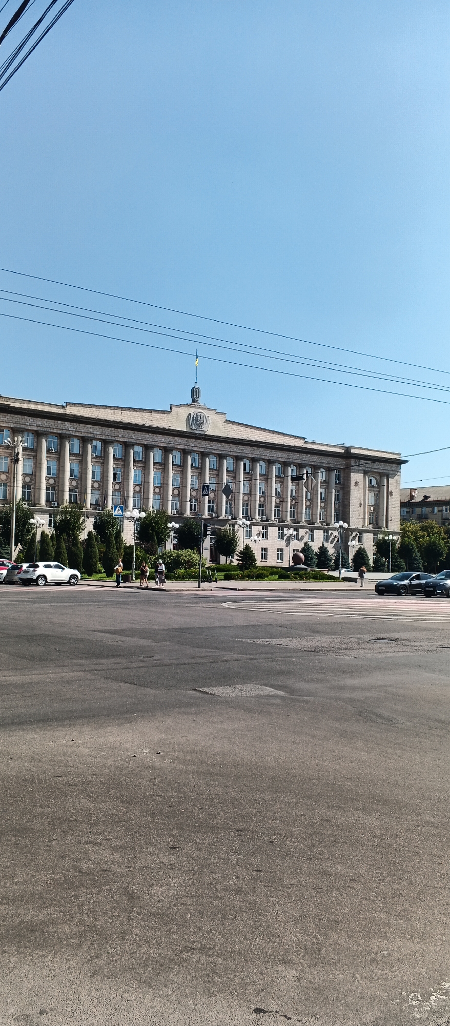 This is a full-shot, eye-level photograph of the Cherkasy Regional State Administration building, located in the city of Cherkasy, Ukraine. The building is a large, multi-story structure with a facade characterized by numerous Ionic columns and evenly spaced windows. At the top center of the building, there is a decorative element topped with a Ukrainian flag. The building is situated behind a green median strip with trees and is visible from across a wide paved intersection, suggesting it is a prominent landmark in the city. The sky is clear and blue. Several cars are present on the road, and a few pedestrians are walking on the sidewalk in front of the building. The foreground of the image shows a section of the asphalt road with markings for lanes and a pedestrian crossing.