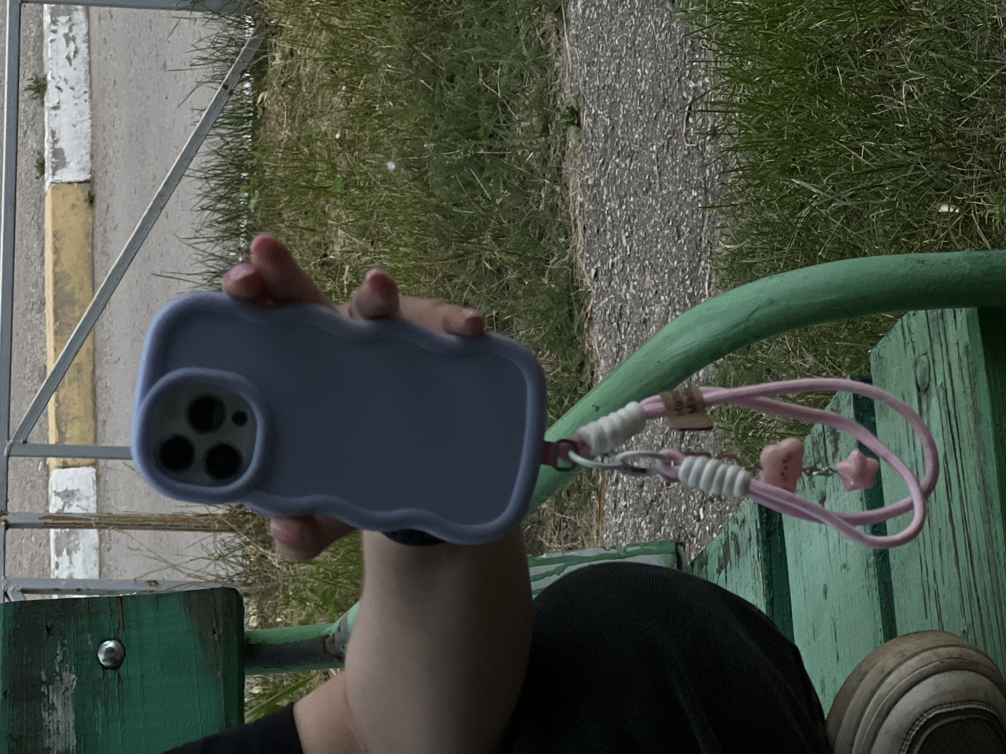 A person, partially visible from the waist down and from the chest up, is sitting on a green wooden bench outdoors. They are holding a light purple phone case with three camera lenses in their right hand. A pink lanyard with small decorative charms, including a pink cloud and a pink star, is attached to the phone case. The person is wearing dark clothing and white and brown shoes. The background consists of a gravel path, green grass, and a metal railing with yellow and white painted concrete barriers behind it. The scene appears to be a public outdoor area, possibly a park or a roadside. No specific landmarks or details are present to pinpoint the location to Ulyanovsk, Russia.