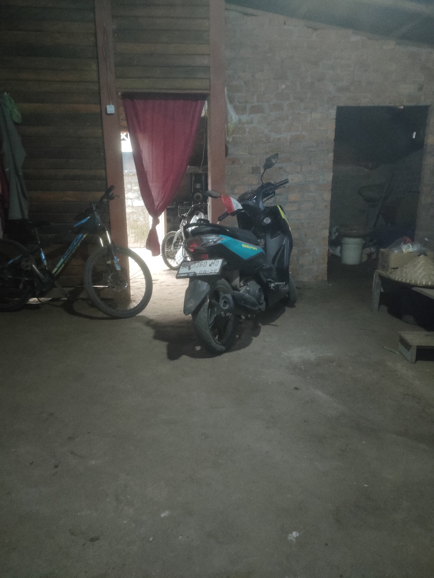 In Padang Dalom, Indonesia, a scene unfolds within what appears to be an unfinished or rustic indoor space. The walls are constructed from rough, unpainted brick on the right and horizontal wooden planks on the left. In the center, a doorway is partially obscured by a draped red cloth, creating a sense of division.
Two motorcycles are the primary subjects. To the right, a scooter-style motorcycle with a blue and black body is parked. Its license plate is visible, reading "BE 3360 HO 08-28". A red and white flag is attached to its handlebars. To the left of the scooter, and positioned further back, is a bicycle with a black frame and blue accents. Its front wheel is turned to the left, and the bike is leaning against a wooden structure.
The floor is concrete and appears somewhat worn. In the background, through the doorway and to the right, more brick walls are visible. Various objects are present in the background, including a white bucket, some boxes or containers, and a woven basket-like item. The lighting is dim, suggesting either evening or an interior space with limited natural light. No people are present in the image.