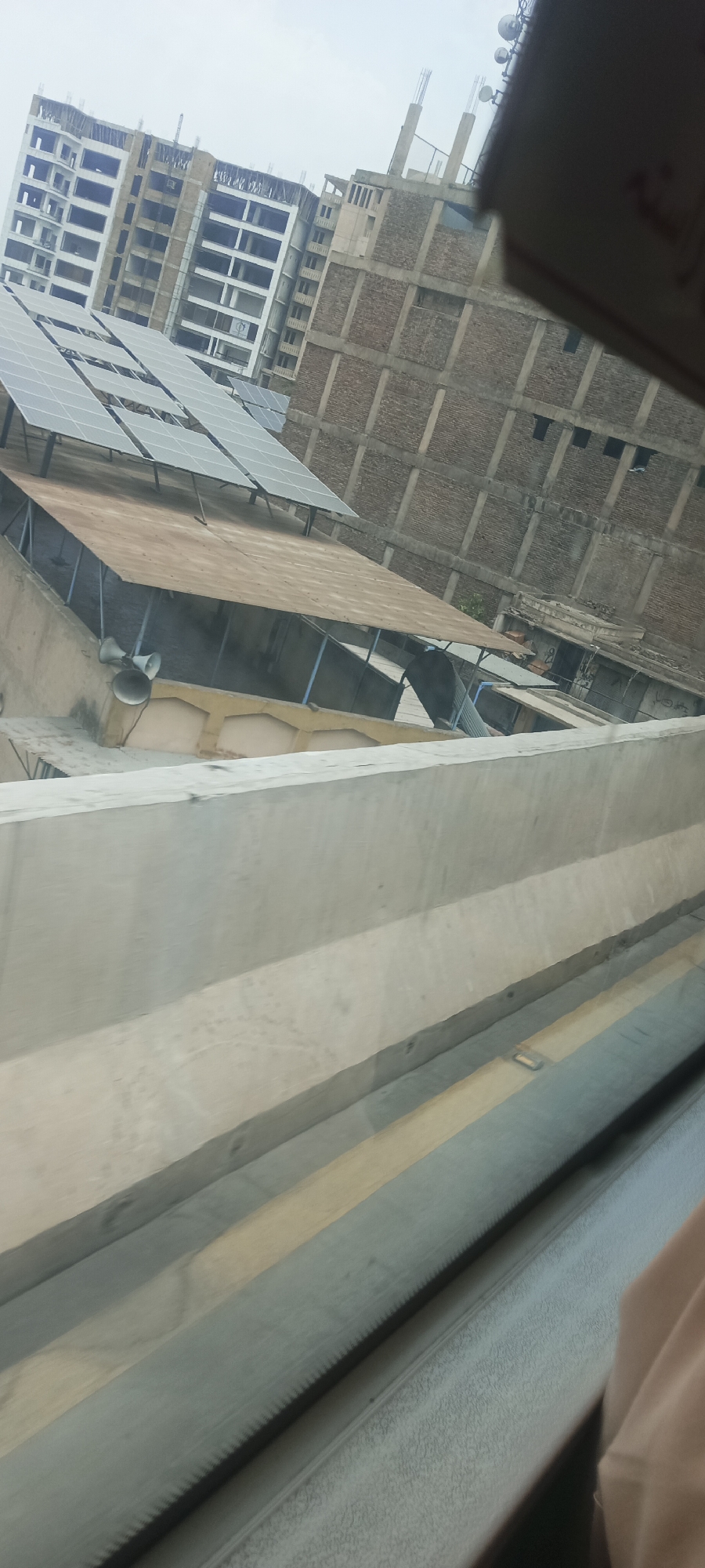 The image depicts a partially constructed building with exposed rebar and brickwork, located in the city of Alam Khan Kalay, Pakistan. Solar panels are installed on a rooftop in the foreground, with a canvas-covered structure underneath. Two loudspeakers are mounted on a wall below the solar panels. The sky is overcast. The perspective suggests the photo was taken from an elevated position, possibly through a window, as a blurred concrete barrier and a dark, ribbed surface are visible in the lower portion of the frame. No people are clearly visible in the image.