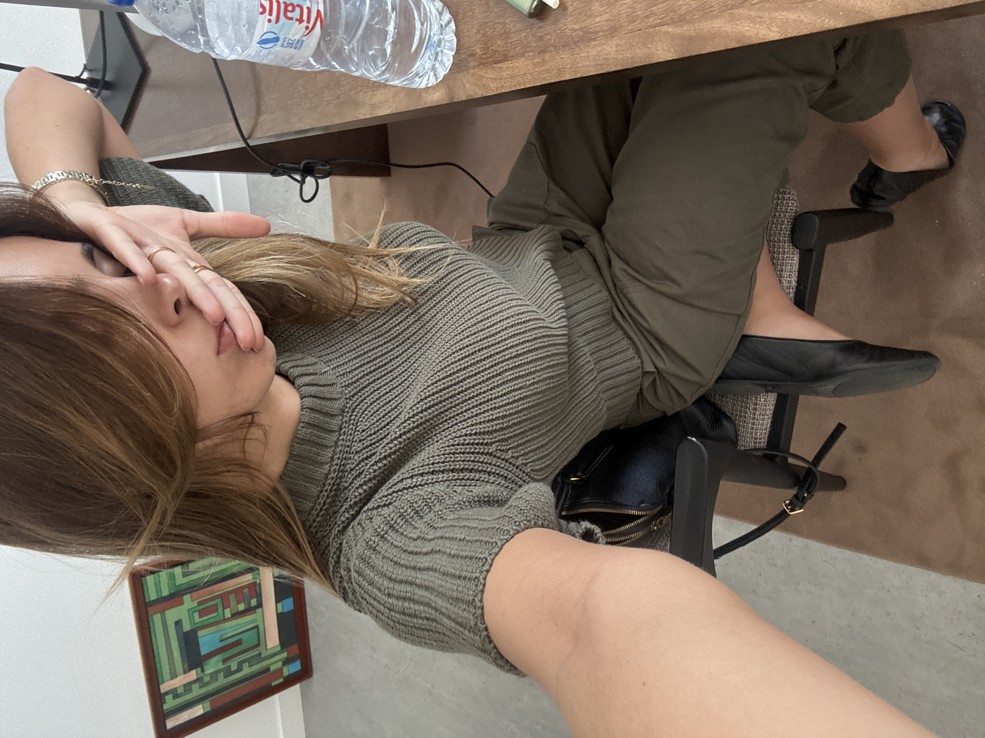 A person is sitting at a desk in a room. The person is wearing an olive green knit sweater and olive green pants, with black flats. There is a clear water bottle on the desk labeled "Vitalis". A black purse is sitting in the chair next to the person. On the wall is a framed piece of art with geometric shapes in green, brown, and blue. The location is likely an indoor space in Torres Vedras, Portugal.