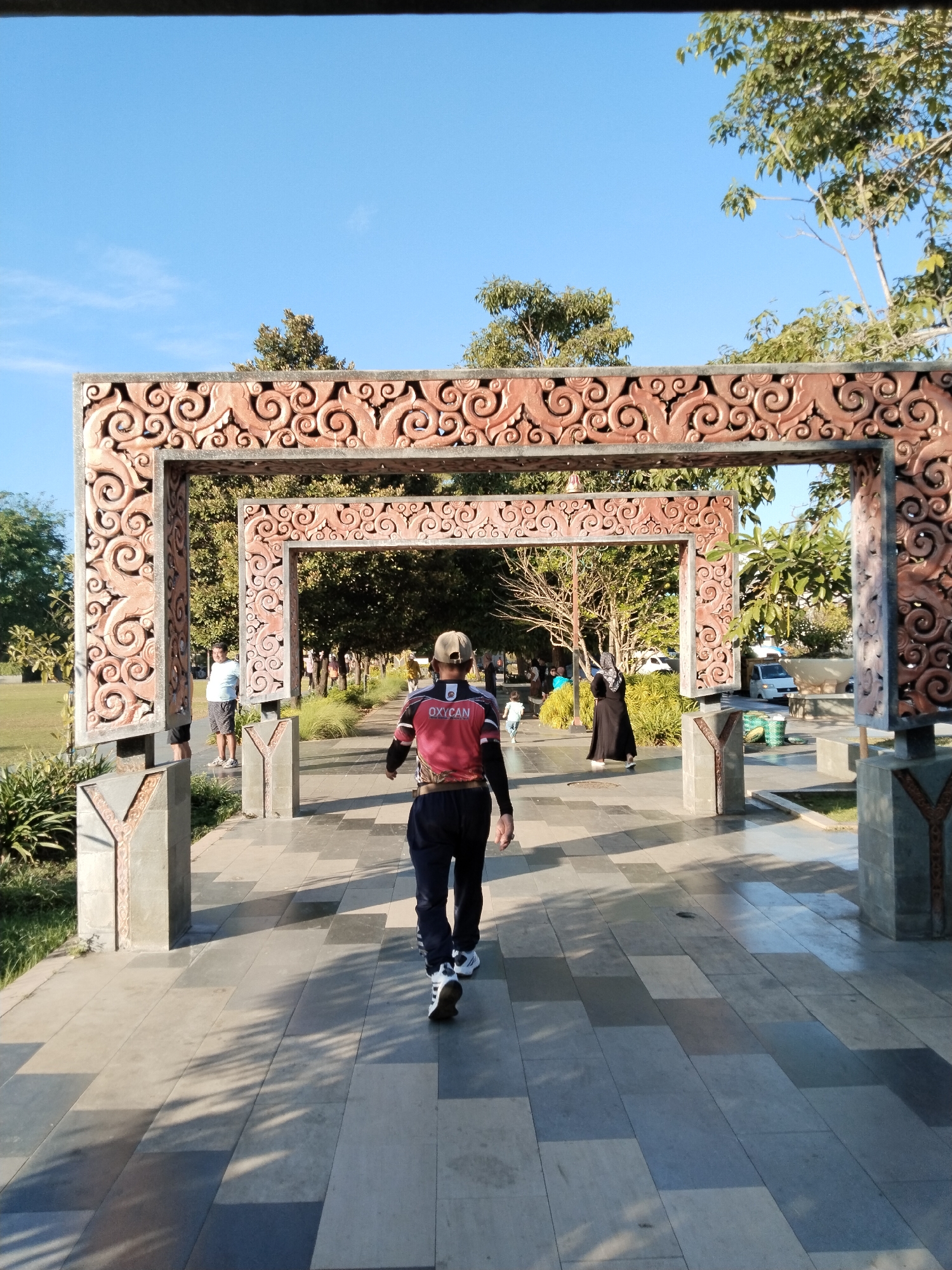 The image shows a man walking away from the camera on a paved pathway lined with ornamental pillars and archways. He is wearing a cap, a red and black shirt with "OXYCAN" visible on the back, and dark pants with white stripes on the sides. He is also wearing white sneakers with black accents. The pathway is paved with large, rectangular tiles of varying shades of gray and light brown, creating a checkered pattern. Sunlight casts shadows from the foliage and the archways onto the path.
In the background, through the archways, several people are visible. To the left, a man in a light blue t-shirt and dark shorts stands near a grassy area. Further back, a woman in a black abaya with a white head covering is walking. Several other figures are also seen in the distance, some appearing to be walking or standing. Vehicles, including a white van, are parked further in the background.
The environment appears to be an outdoor park or public space, with trees and lush green grass visible on either side of the pathway. The sky is a clear, bright blue. The ornamental archways are made of a material that appears to be carved wood or a similar material with intricate swirling patterns. The pillars supporting the archways are gray and rectangular, with decorative Y-shaped elements at the base. The overall scene suggests a pleasant, sunny day in a well-maintained outdoor area. The location is identified as Kecamatan Pati, Indonesia.