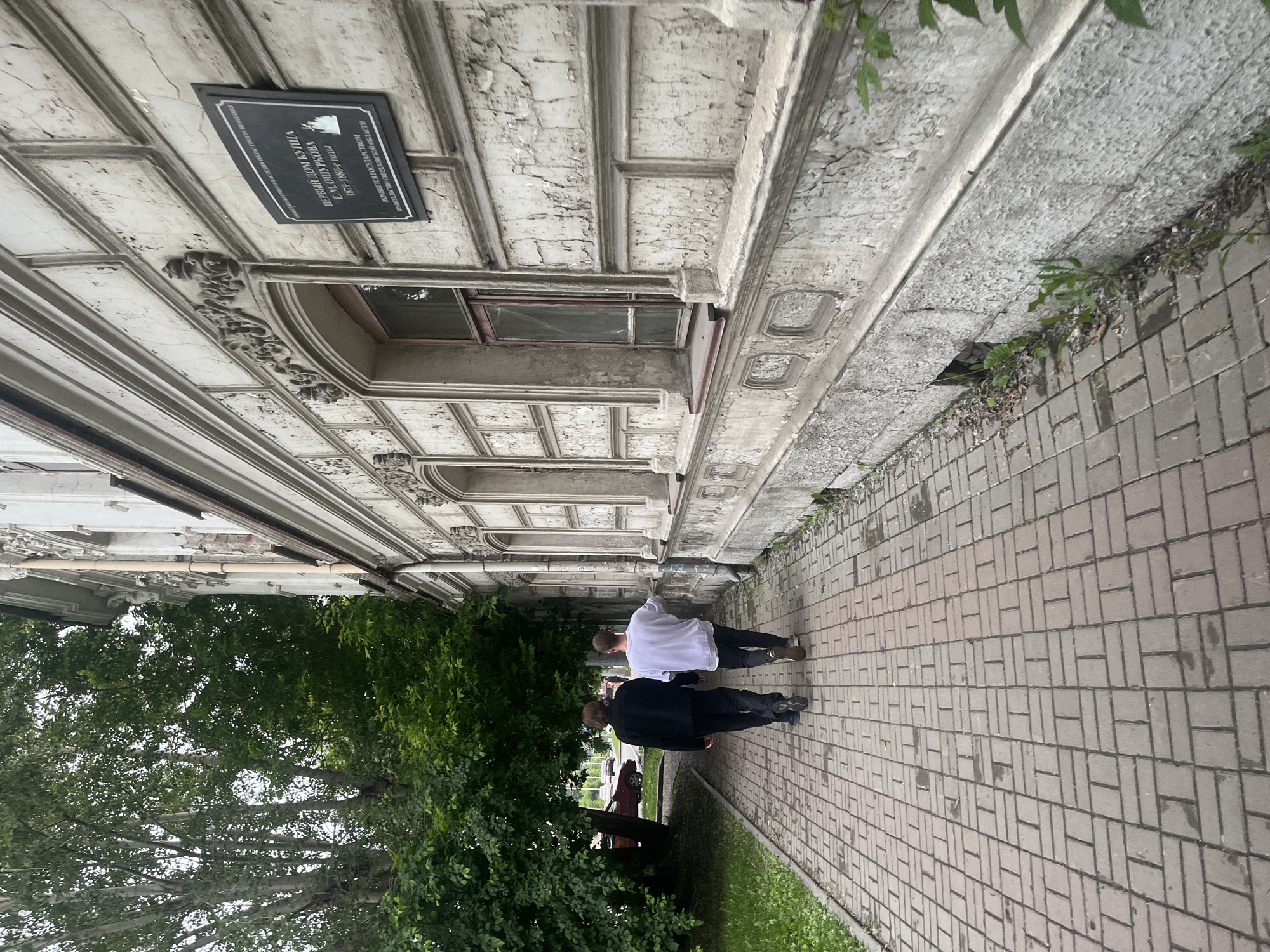 Two individuals walk away from the camera on a brick-paved sidewalk adjacent to a weathered, ornate building. The person on the left wears a black jacket and dark pants, while the person on the right wears a white long-sleeved shirt and dark pants. They are walking towards the left of the frame. The building's facade features textured walls with peeling paint, decorative moldings, and arched windows, one of which is partially visible and has a wooden frame. A dark plaque with text in a foreign language is mounted on the building's wall. To the left of the sidewalk, lush green trees with a dense canopy are present, partially obscuring a red car visible in the background. The ground appears damp in patches on the sidewalk and is bordered by green grass along the building's edge. The scene suggests an overcast or shaded environment.