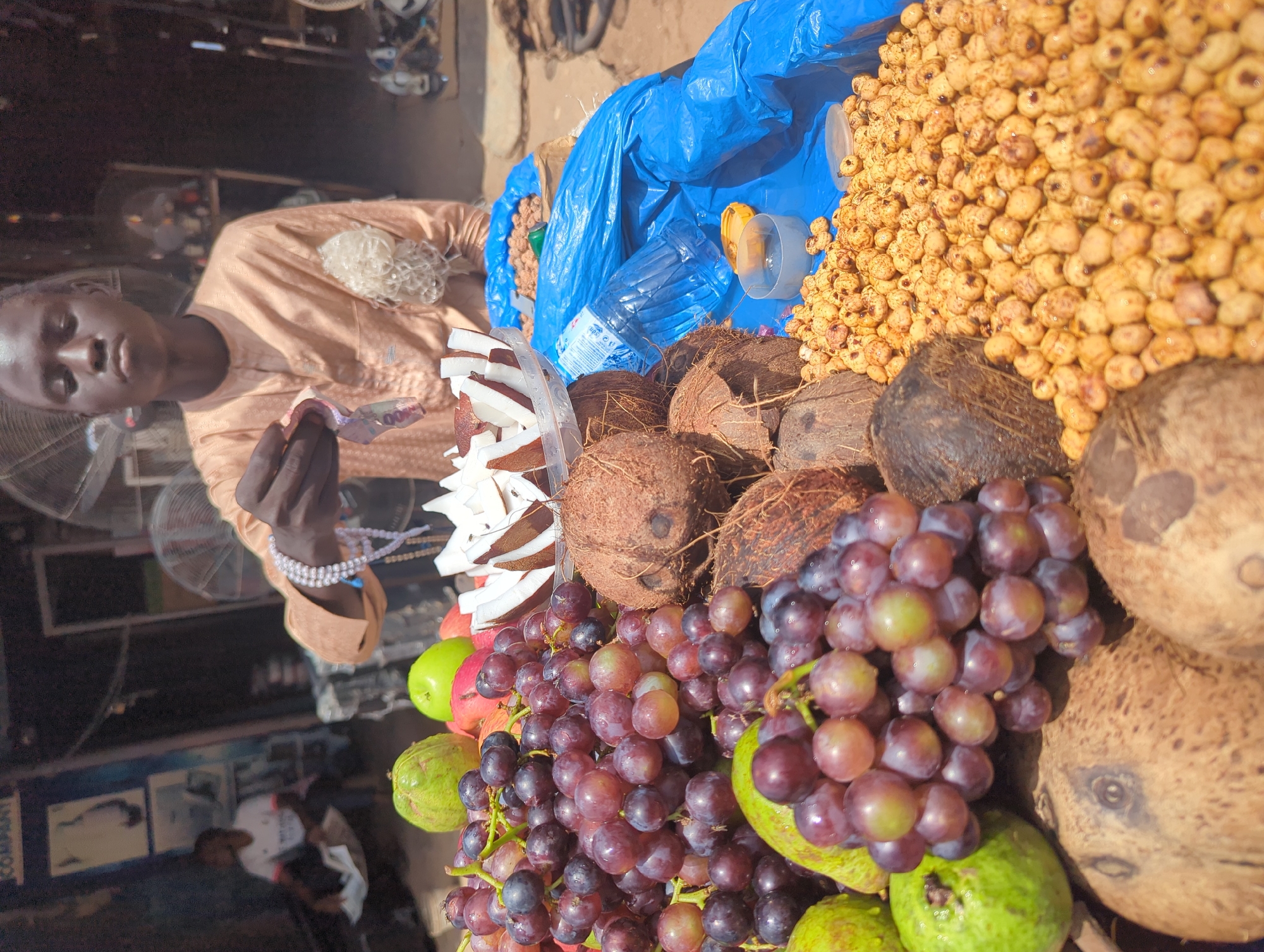 A young man, dressed in a tan, long-sleeved tunic, stands behind a market stall in Shaba, Nigeria. He is holding out what appears to be Nigerian currency in his right hand. In front of him is an assortment of fruits and nuts. Bunches of red grapes, several green guavas, and two green apples are visible. Slices of coconut are arranged in a clear plastic bowl. Several whole coconuts are on the table, along with a pile of small, round, light brown nuts. A blue plastic sheet covers part of the stall, and a clear plastic bottle of water rests on top of it. In the background, another person is seated, and part of a fan can be seen. The scene suggests a bustling outdoor market environment.