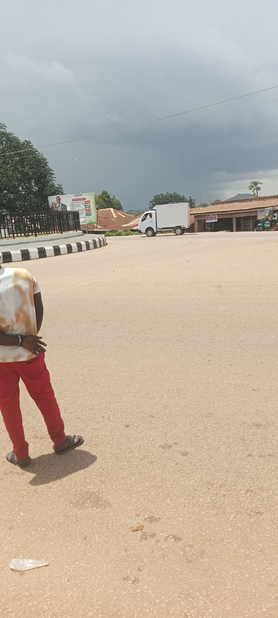 The image depicts a street scene in Kuru Dungus, Nigeria. In the foreground, the lower body of a person wearing a tie-dye t-shirt and red pants is visible, facing away from the camera and walking on an asphalt road. The person is wearing sandals. In the mid-ground, a white refrigerated truck is driving on the road, and a small green tricycle with a rider is parked to the right. Behind the truck, buildings with brown roofs are visible. To the left, a billboard with images of a man and a woman is displayed, and behind it are trees. The road is paved with asphalt and has a curb with black and white stripes. The sky is overcast with dark clouds, suggesting potential rain. A discarded plastic bottle is on the ground in the lower left corner.